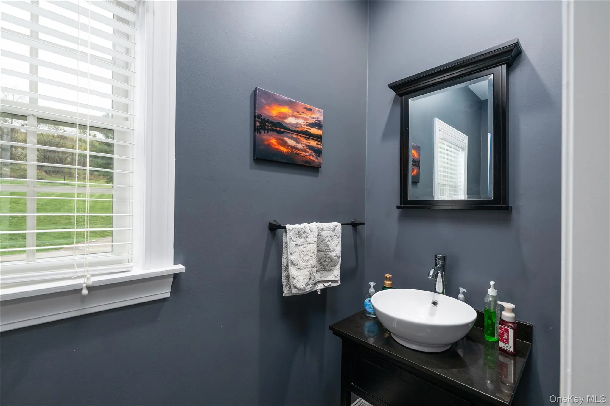 First floor Powder room featuring a single window with blinds, a wall-mounted mirror with a black frame, a dark wood-finish vanity with a vessel sink, and a chrome faucet First floor Powder room featuring a single window with blinds, a wall-mounted mirror with a black frame, a dark wood-finish vanity with a vessel sink, and a chrome faucet