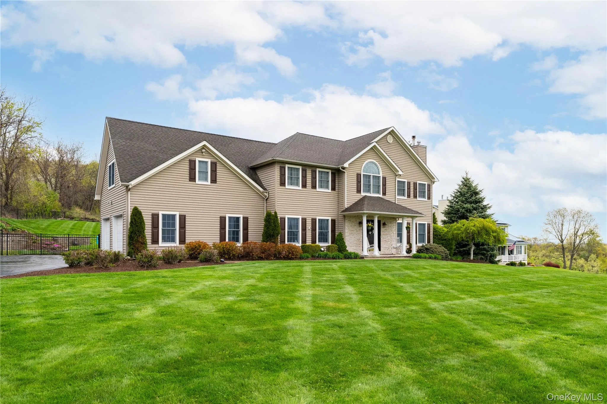 Spacious suburban residence featuring a tan siding facade with dark shutters, a built-in multi-car garage, a covered front entry with stone accents, a large manicured lawn, and mature landscaping Spacious suburban residence featuring a tan siding facade with dark shutters, a built-in multi-car garage, a covered front entry with stone accents, a large manicured lawn, and mature landscaping