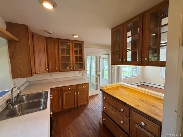 Kitchen featuring wood cabinetry, some with glass-front doors, a double basin stainless steel sink, light-toned countertops, and wood-finish flooring Kitchen featuring wood cabinetry, some with glass-front doors, a double basin stainless steel sink, light-toned countertops, and wood-finish flooring