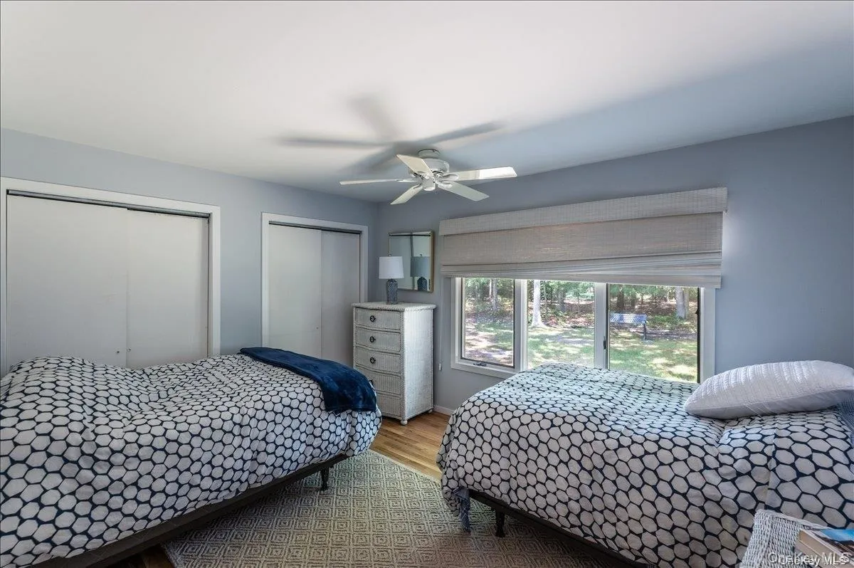 Bedroom featuring wood-finish flooring, two sliding door closets, a ceiling fan, and a large window overlooking the exterior landscape Bedroom featuring wood-finish flooring, two sliding door closets, a ceiling fan, and a large window overlooking the exterior landscape