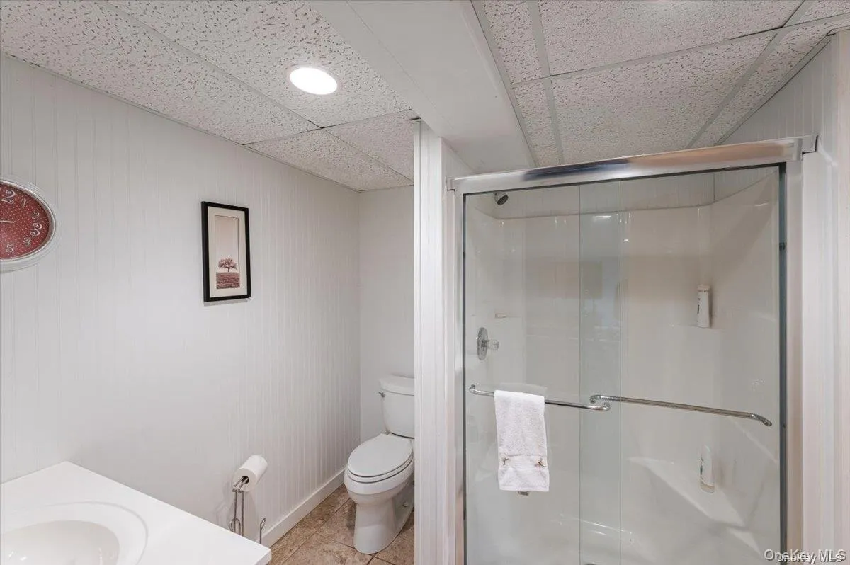 Bathroom featuring a glass-enclosed shower, white vanity with integrated sink, toilet, textured ceiling, and vertical paneling on walls Bathroom featuring a glass-enclosed shower, white vanity with integrated sink, toilet, textured ceiling, and vertical paneling on walls