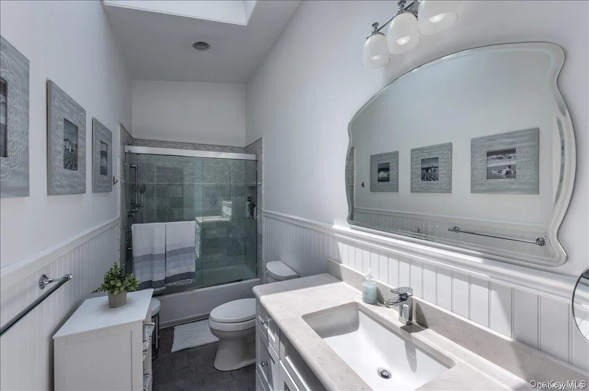 Bathroom featuring a white vanity with an integrated sink and light-toned countertop, a frameless shower-tub combination with sliding glass doors, white beadboard wainscoting, and a skylight Bathroom featuring a white vanity with an integrated sink and light-toned countertop, a frameless shower-tub combination with sliding glass doors, white beadboard wainscoting, and a skylight