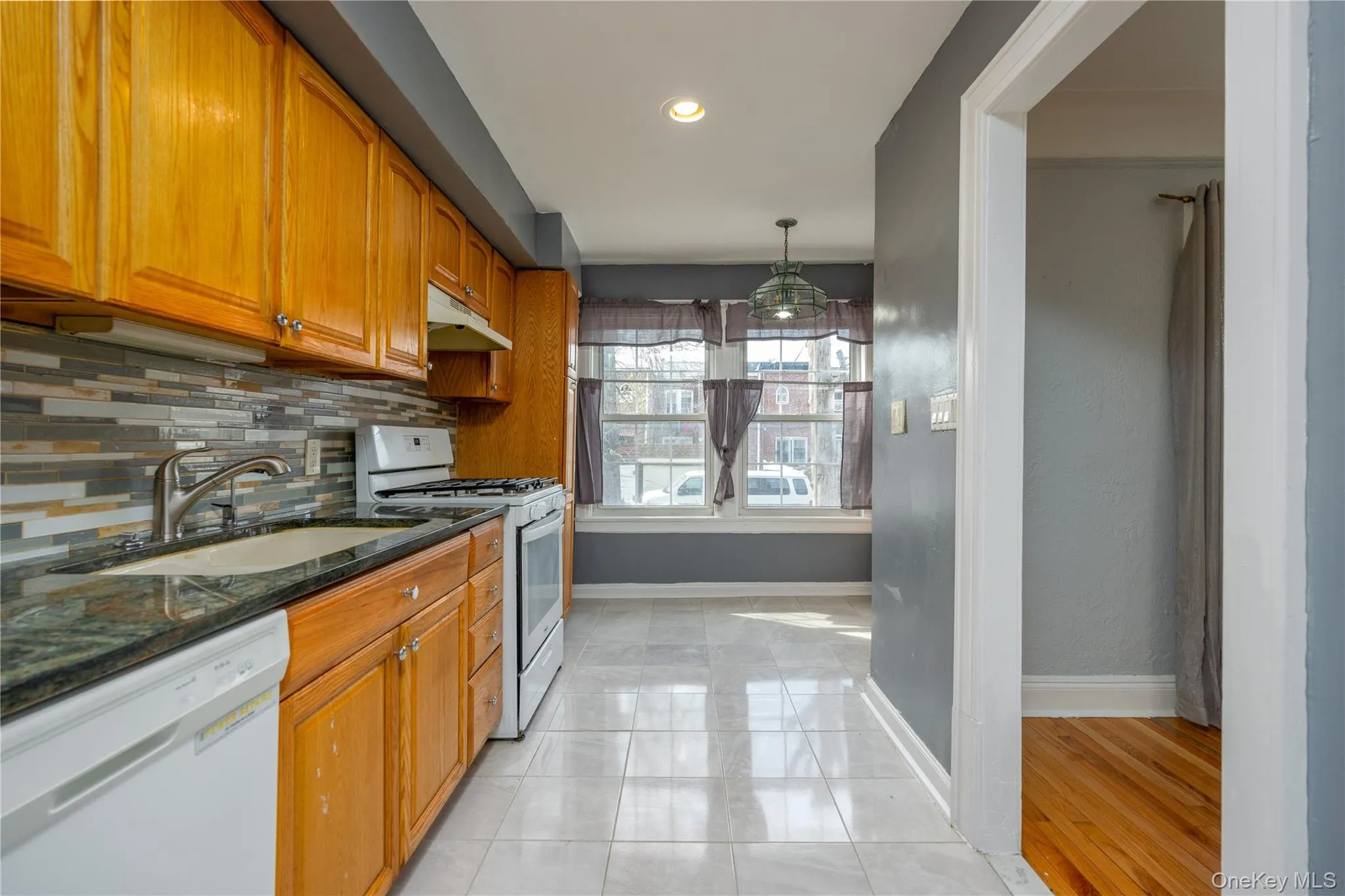 Galley kitchen 9'5" x 14'11"Kitchen featuring wood cabinetry, a mosaic tile backsplash, dark countertops, white appliances, and light-colored tile flooring Galley kitchen 9'5" x 14'11"Kitchen featuring wood cabinetry, a mosaic tile backsplash, dark countertops, white appliances, and light-colored tile flooring