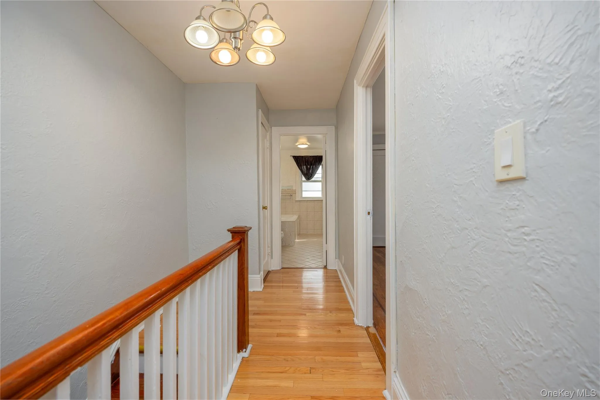Second floor hallway featuring wood-finish flooring, white painted balusters, a wood-finish handrail, textured light gray walls, and a five-light ceiling fixture Second floor hallway featuring wood-finish flooring, white painted balusters, a wood-finish handrail, textured light gray walls, and a five-light ceiling fixture