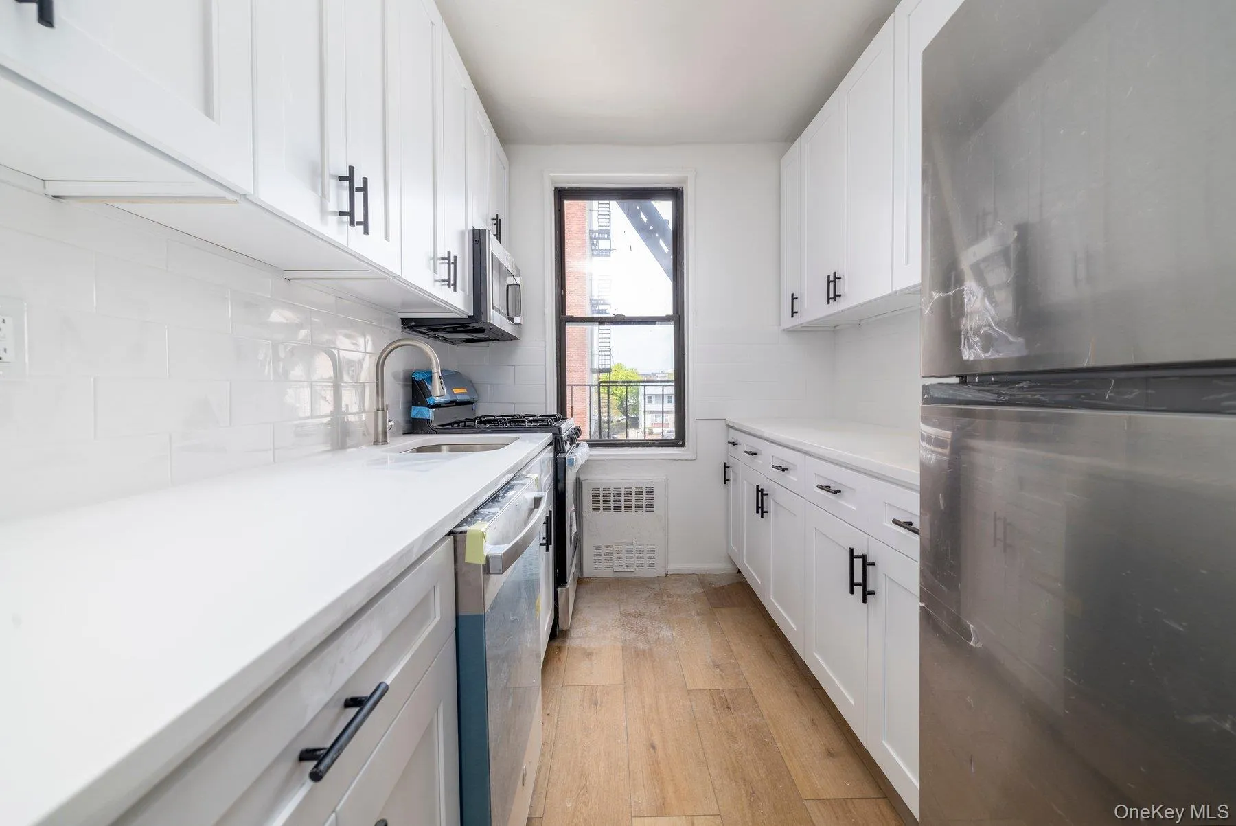 Bright galley kitchen featuring white shaker cabinetry, light countertops, wood-finish flooring, a stainless steel refrigerator, and a window providing natural light Bright galley kitchen featuring white shaker cabinetry, light countertops, wood-finish flooring, a stainless steel refrigerator, and a window providing natural light