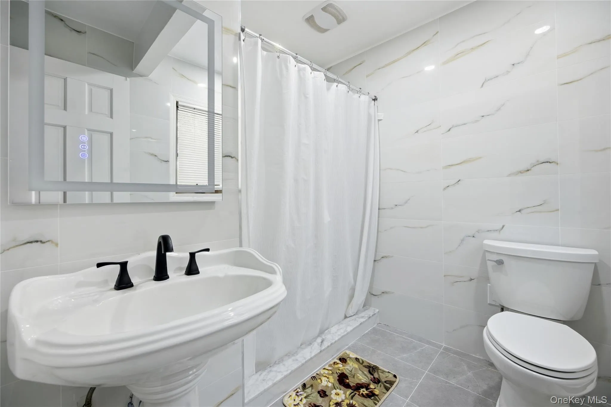 Pedestal sink with matte black faucet, illuminated mirror, white tiled walls with subtle veining, and grey tiled flooring Pedestal sink with matte black faucet, illuminated mirror, white tiled walls with subtle veining, and grey tiled flooring