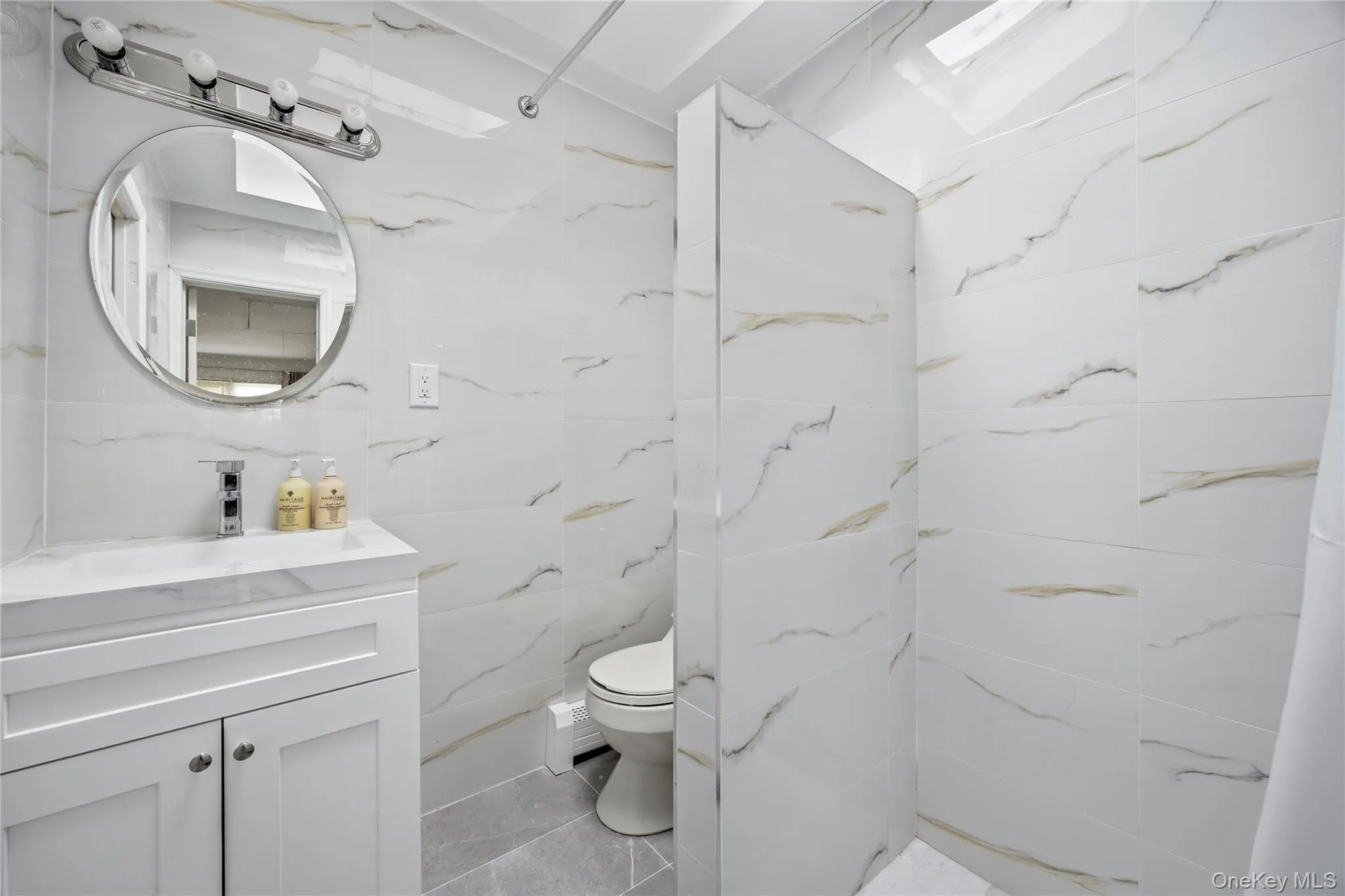 Bathroom featuring a white vanity with an integrated sink, round chrome-framed mirror, and chrome vanity lighting Bathroom featuring a white vanity with an integrated sink, round chrome-framed mirror, and chrome vanity lighting