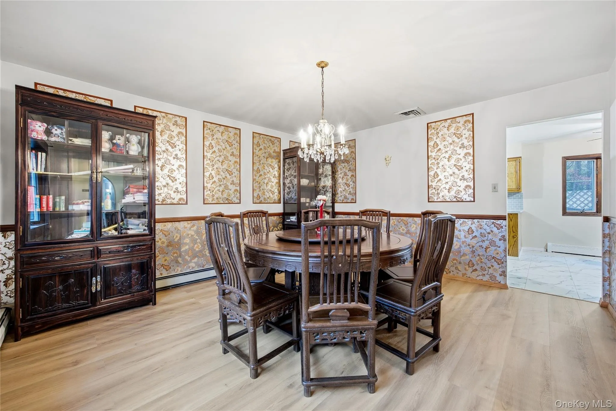 Dining area featuring wood-finish flooring, a tiered chandelier, decorative wall panels, and an open doorway to an adjacent space Dining area featuring wood-finish flooring, a tiered chandelier, decorative wall panels, and an open doorway to an adjacent space