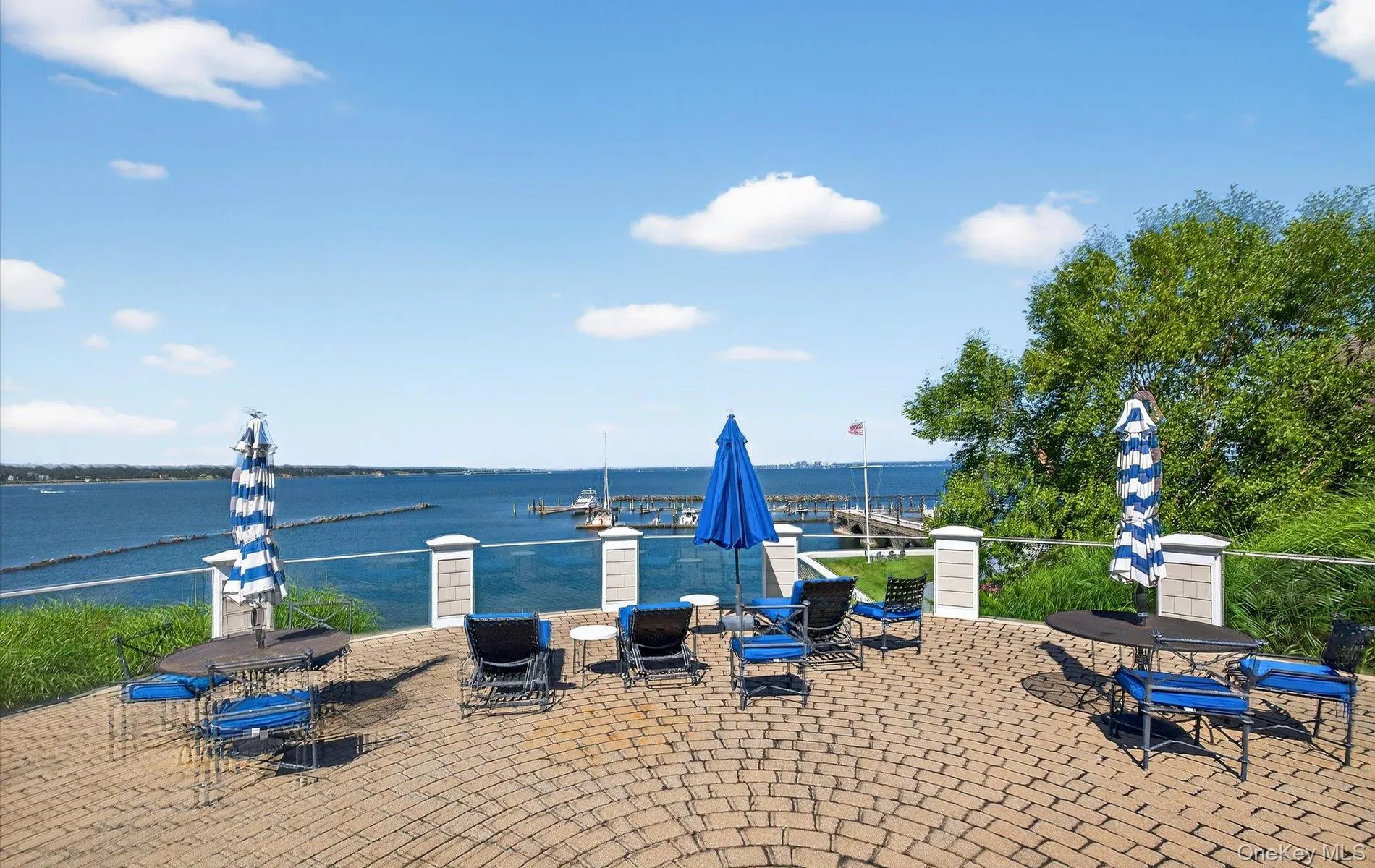 Expansive brick paver patio featuring a clear panel railing with decorative posts, overlooking a body of water with a distant marina Expansive brick paver patio featuring a clear panel railing with decorative posts, overlooking a body of water with a distant marina