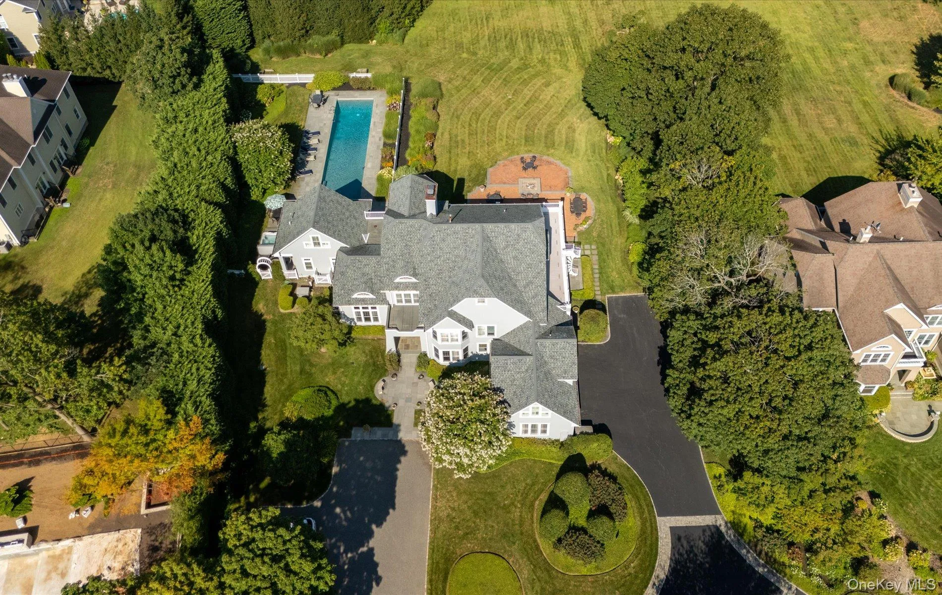 Expansive property featuring a multi-level residence with a gray shingle roof, a rectangular in-ground pool, a circular patio, mature perimeter plantings, and a paved driveway Expansive property featuring a multi-level residence with a gray shingle roof, a rectangular in-ground pool, a circular patio, mature perimeter plantings, and a paved driveway