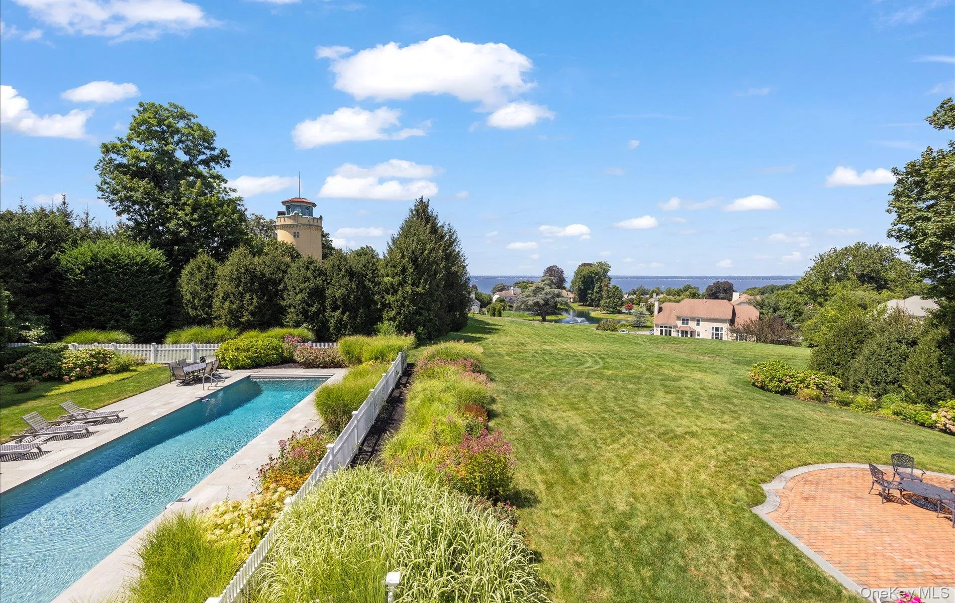 Elongated swimming pool with surrounding hardscaping, a large manicured lawn, a circular paver patio, and mature trees and landscaping Elongated swimming pool with surrounding hardscaping, a large manicured lawn, a circular paver patio, and mature trees and landscaping