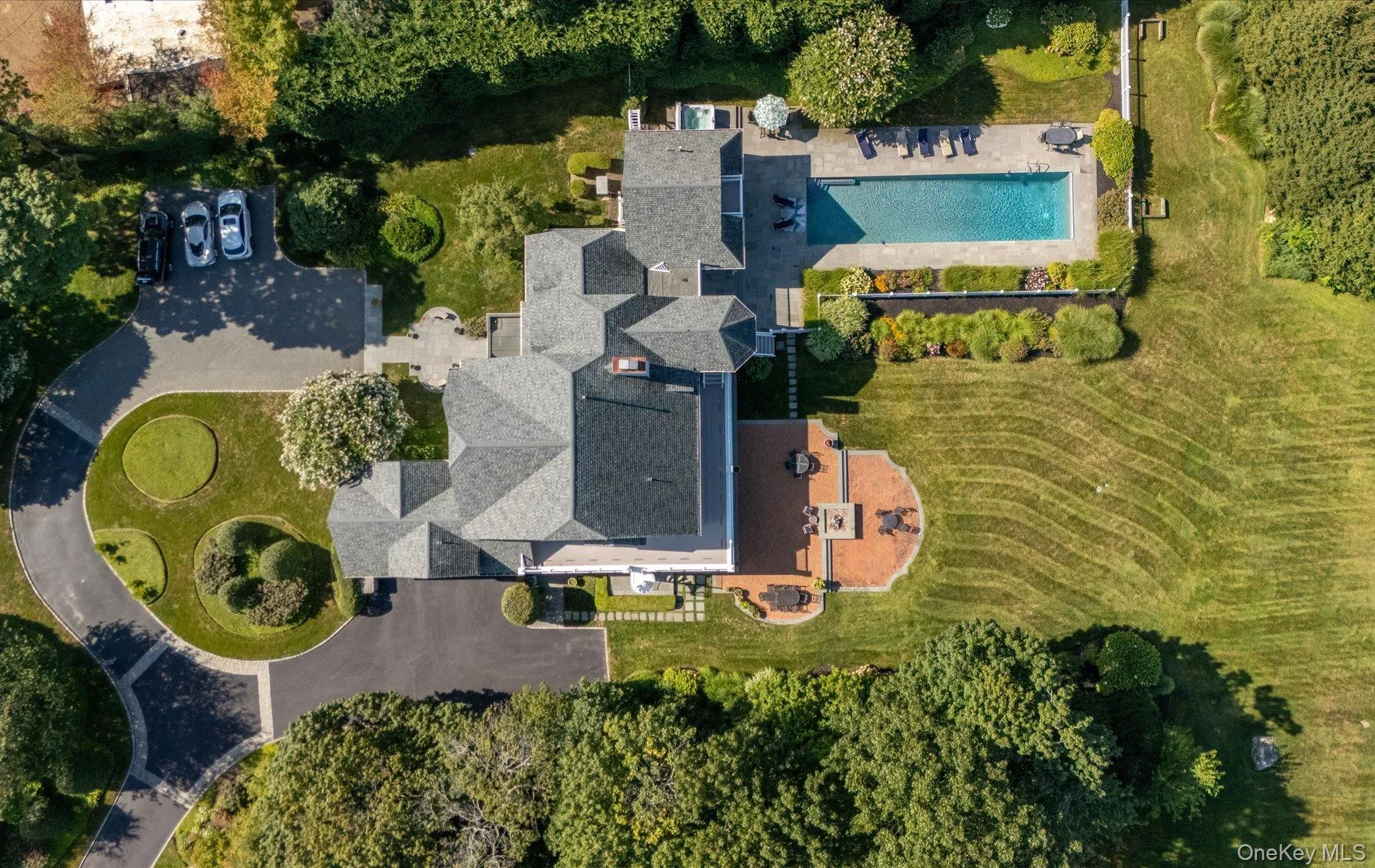 Extensive property featuring a multi-gabled main residence, a large swimming pool with surrounding patio, a curved driveway with landscaped islands, a spacious brick patio, and a broad lawn area Extensive property featuring a multi-gabled main residence, a large swimming pool with surrounding patio, a curved driveway with landscaped islands, a spacious brick patio, and a broad lawn area