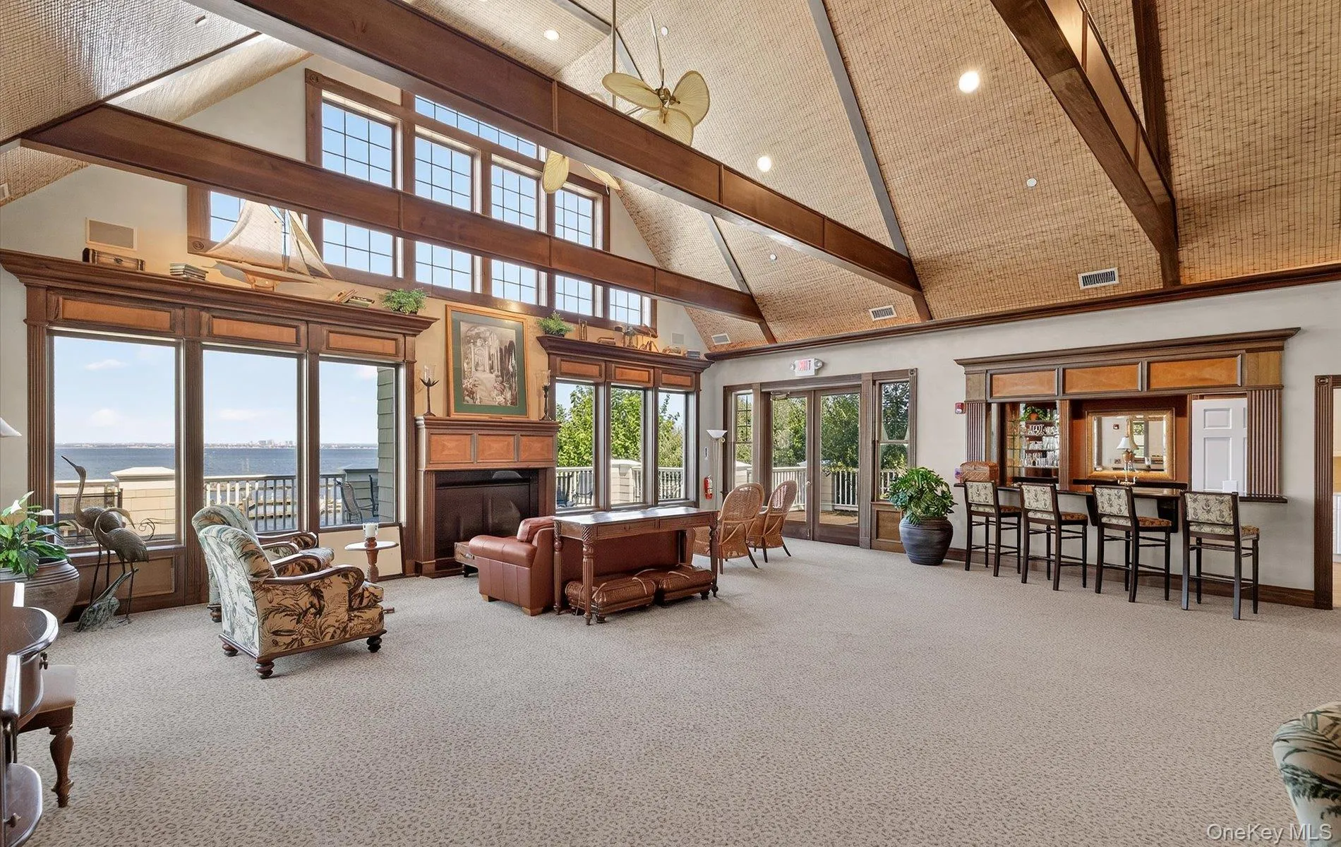 Expansive great room featuring a vaulted ceiling with exposed wood beams, a built-in wet bar, a fireplace with a wood mantle, and floor-to-ceiling windows overlooking the water Expansive great room featuring a vaulted ceiling with exposed wood beams, a built-in wet bar, a fireplace with a wood mantle, and floor-to-ceiling windows overlooking the water