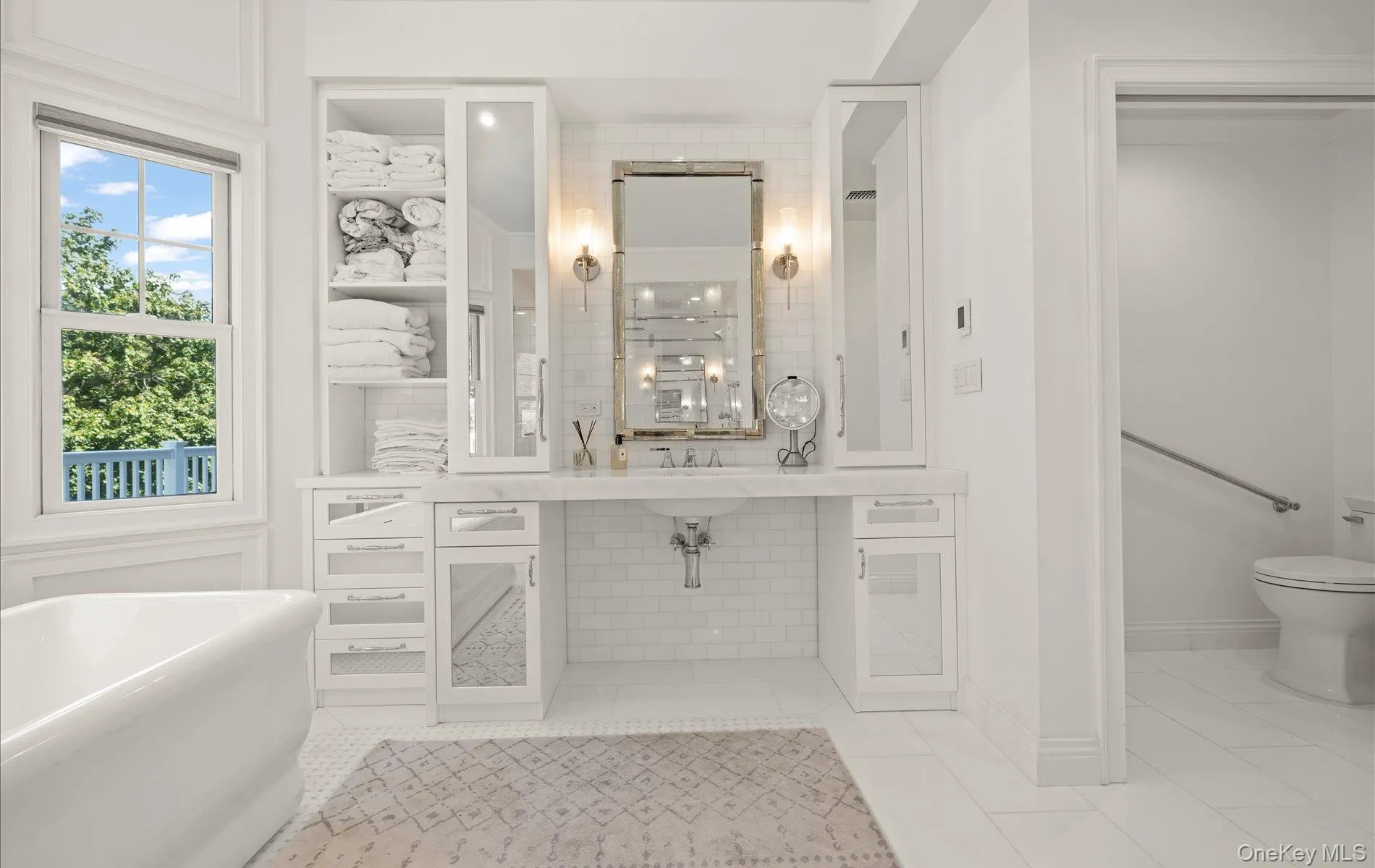 White bathroom featuring a freestanding soaking tub, a vanity with a white countertop and undermount sink, built-in storage cabinetry, a framed mirror with wall sconces, and white subway tile backsplash White bathroom featuring a freestanding soaking tub, a vanity with a white countertop and undermount sink, built-in storage cabinetry, a framed mirror with wall sconces, and white subway tile backsplash
