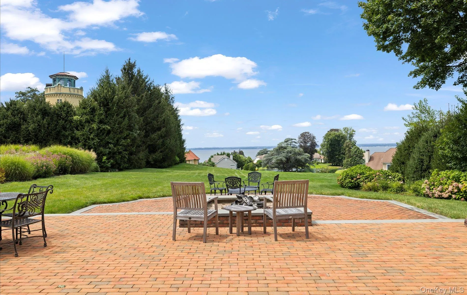 Expansive brick paver patio featuring a fire pit area, surrounded by a manicured lawn and mature landscaping Expansive brick paver patio featuring a fire pit area, surrounded by a manicured lawn and mature landscaping