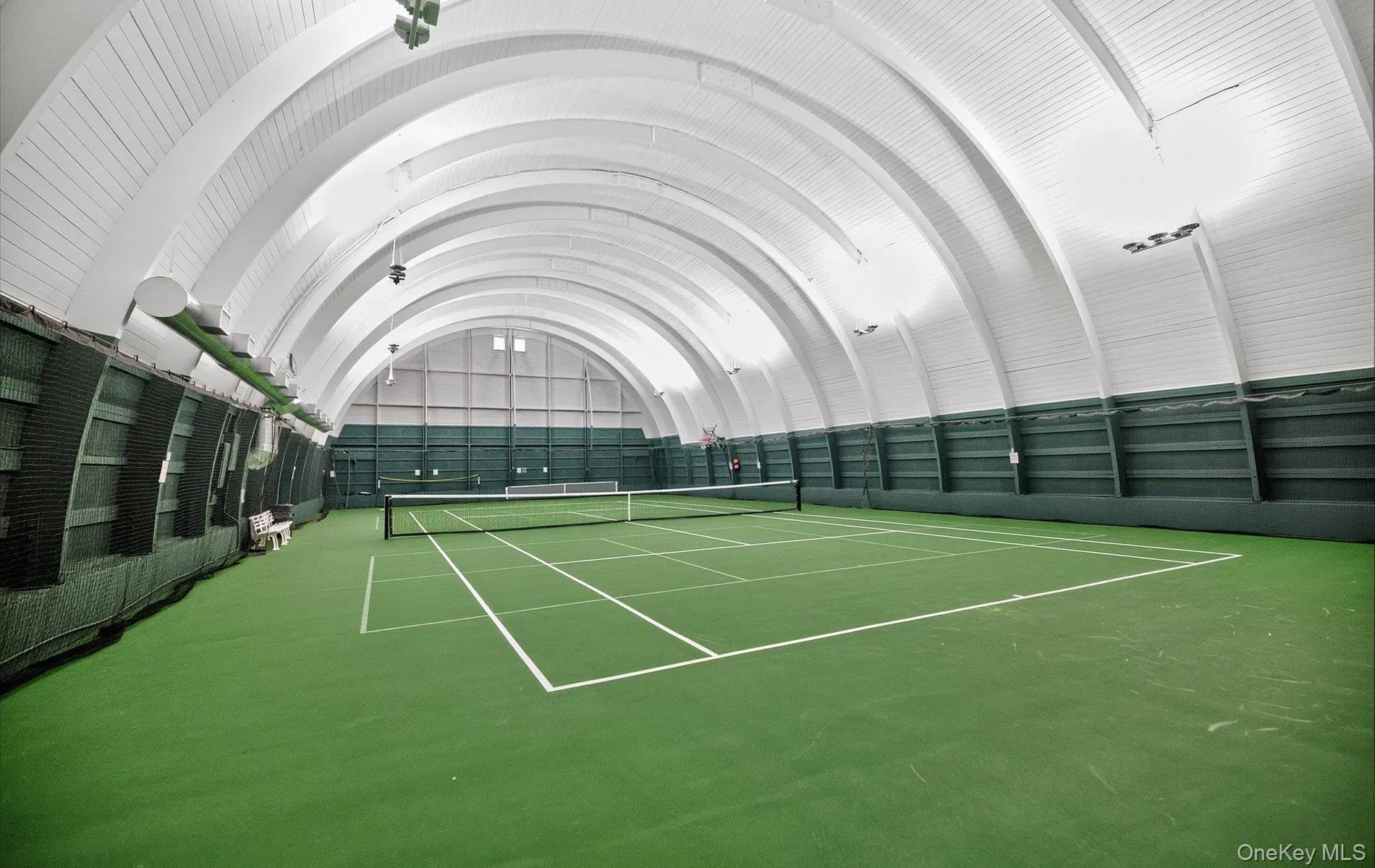 Indoor tennis court featuring a green playing surface with white line markings, an arched ceiling structure, integrated lighting fixtures, and a protective netting system along the side walls Indoor tennis court featuring a green playing surface with white line markings, an arched ceiling structure, integrated lighting fixtures, and a protective netting system along the side walls