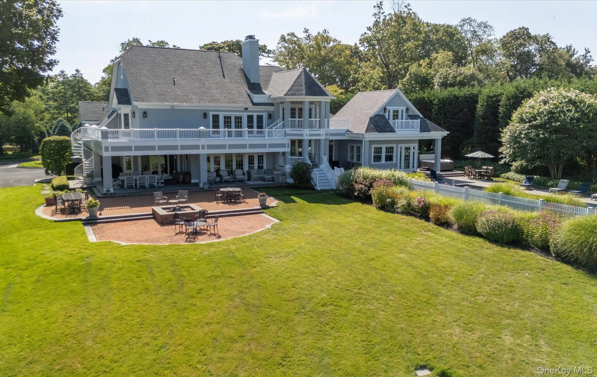 Expansive rear facade featuring a second-story balcony with a spiral staircase, multiple outdoor patio areas, a built-in fire pit, and extensive green space bordered by mature landscaping Expansive rear facade featuring a second-story balcony with a spiral staircase, multiple outdoor patio areas, a built-in fire pit, and extensive green space bordered by mature landscaping