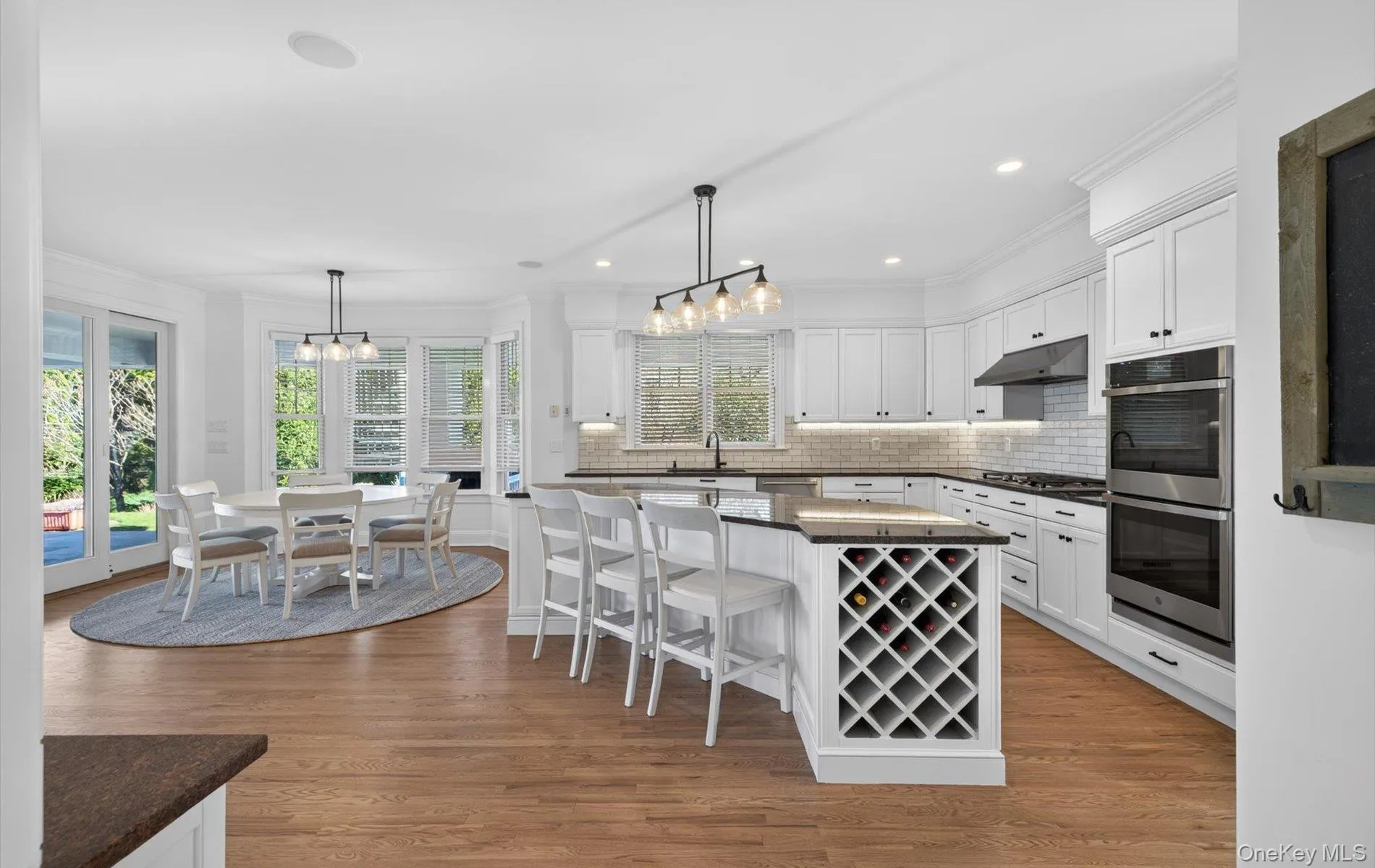 Kitchen featuring white cabinetry, wood-finish flooring, a central island with integrated wine storage, stainless steel wall ovens, and a subway tile backsplash Kitchen featuring white cabinetry, wood-finish flooring, a central island with integrated wine storage, stainless steel wall ovens, and a subway tile backsplash