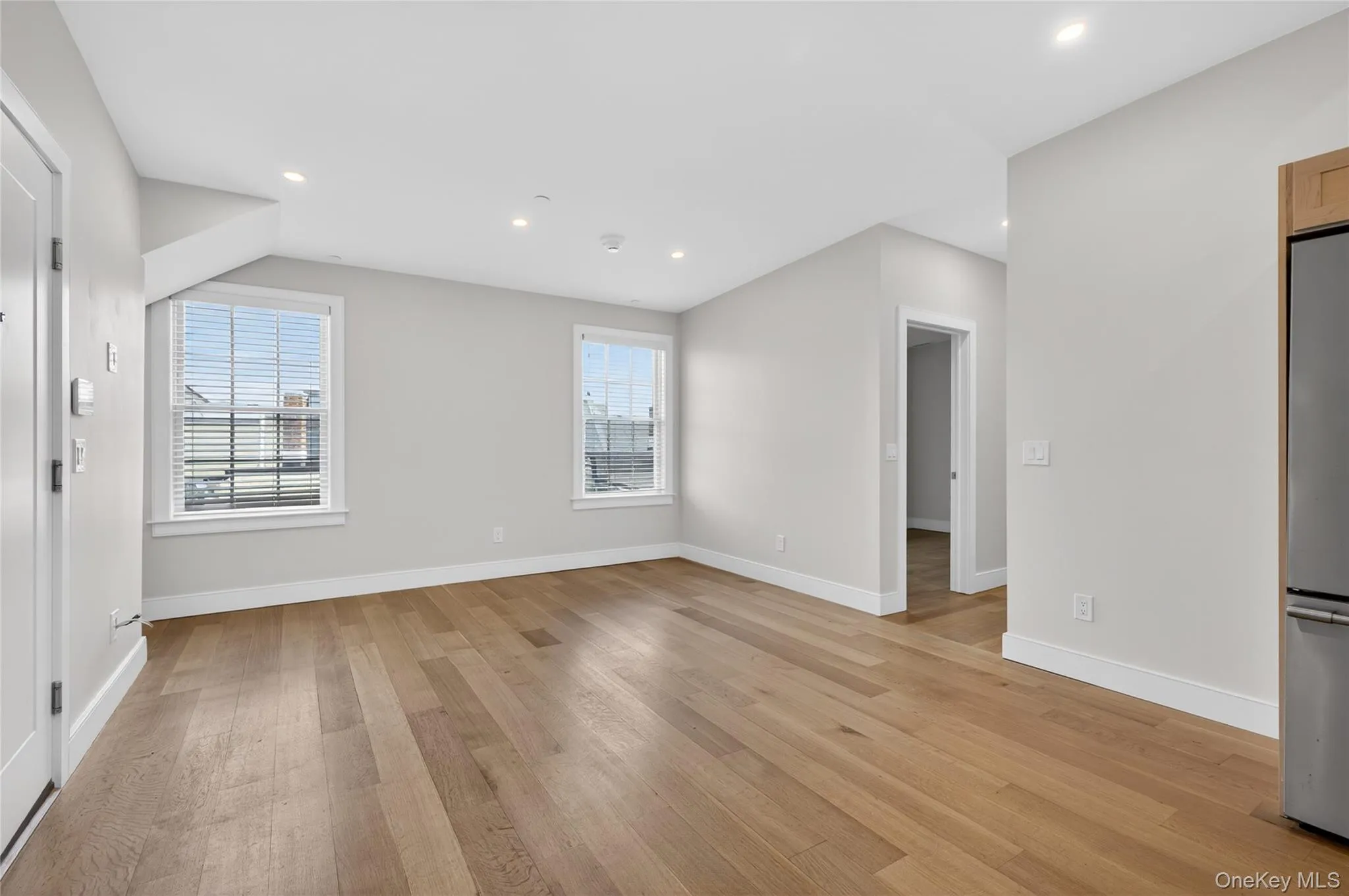 Spacious living area featuring wood-finish flooring, two windows with blinds, recessed ceiling lighting, and a stainless steel refrigerator Spacious living area featuring wood-finish flooring, two windows with blinds, recessed ceiling lighting, and a stainless steel refrigerator