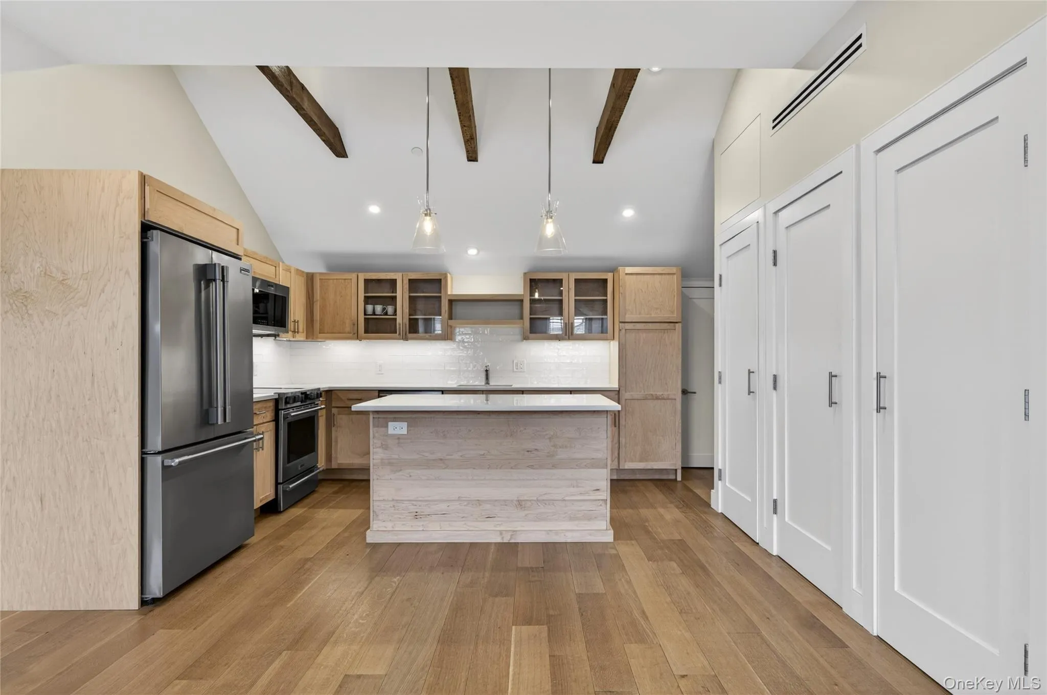 Modern kitchen featuring a central island, stainless steel appliances, wood-finish flooring, exposed ceiling beams, and integrated white cabinetry Modern kitchen featuring a central island, stainless steel appliances, wood-finish flooring, exposed ceiling beams, and integrated white cabinetry