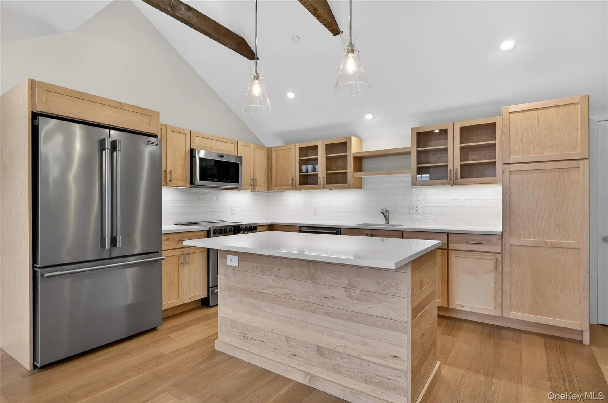 Modern kitchen featuring a vaulted ceiling with exposed wood beams, light wood-finish cabinetry, stainless steel appliances, a white subway tile backsplash, and a central island with a light-toned quartz countertop Modern kitchen featuring a vaulted ceiling with exposed wood beams, light wood-finish cabinetry, stainless steel appliances, a white subway tile backsplash, and a central island with a light-toned quartz countertop