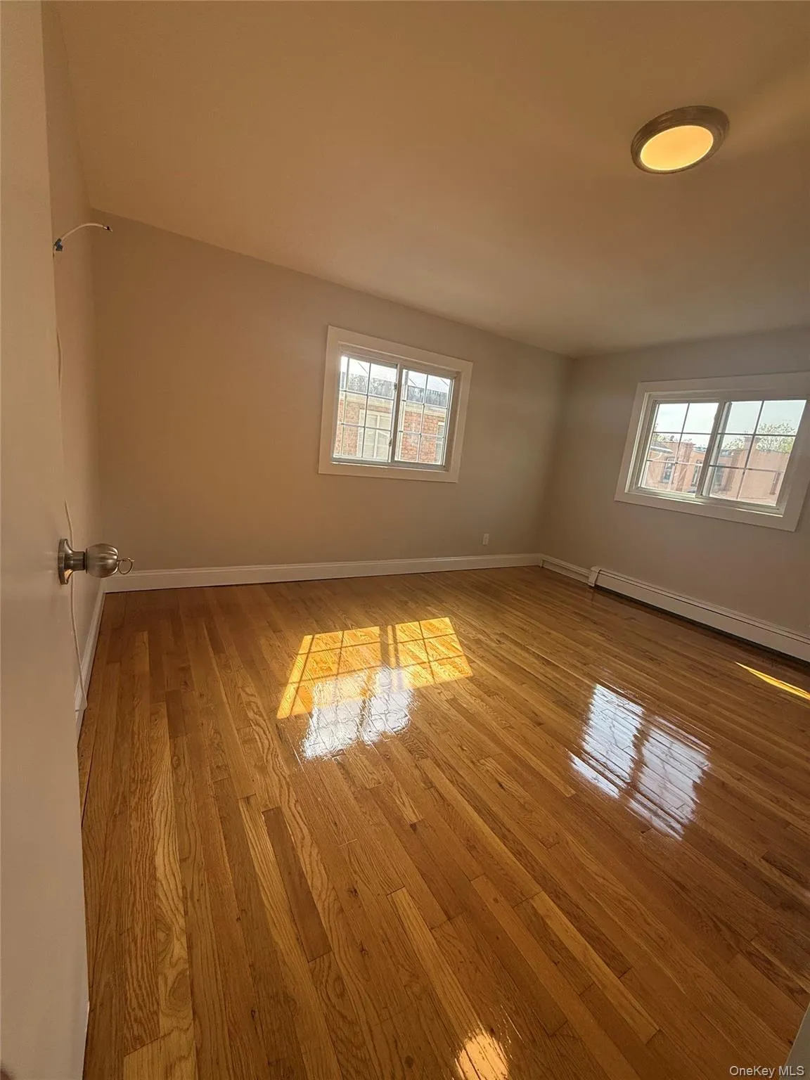 Room featuring wood flooring, two windows, baseboard heating, a flush-mount ceiling light, and light-toned wall paint Room featuring wood flooring, two windows, baseboard heating, a flush-mount ceiling light, and light-toned wall paint