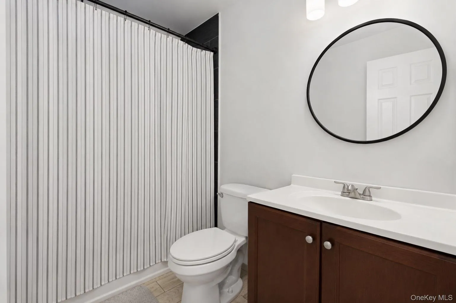 Bathroom featuring a dark wood-finish vanity with a white countertop and integrated sink, a large round black-framed mirror, and a contemporary vanity light fixture Bathroom featuring a dark wood-finish vanity with a white countertop and integrated sink, a large round black-framed mirror, and a contemporary vanity light fixture
