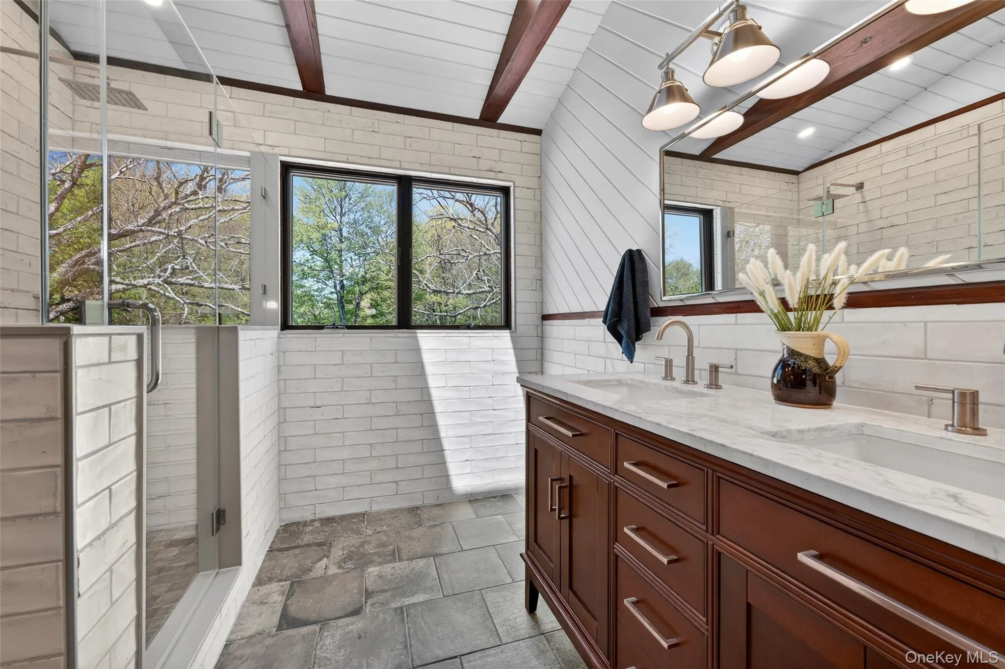 The spa-like primary en suite features distressed subway tile, open ceilings with wood beams and double sinks The spa-like primary en suite features distressed subway tile, open ceilings with wood beams and double sinks