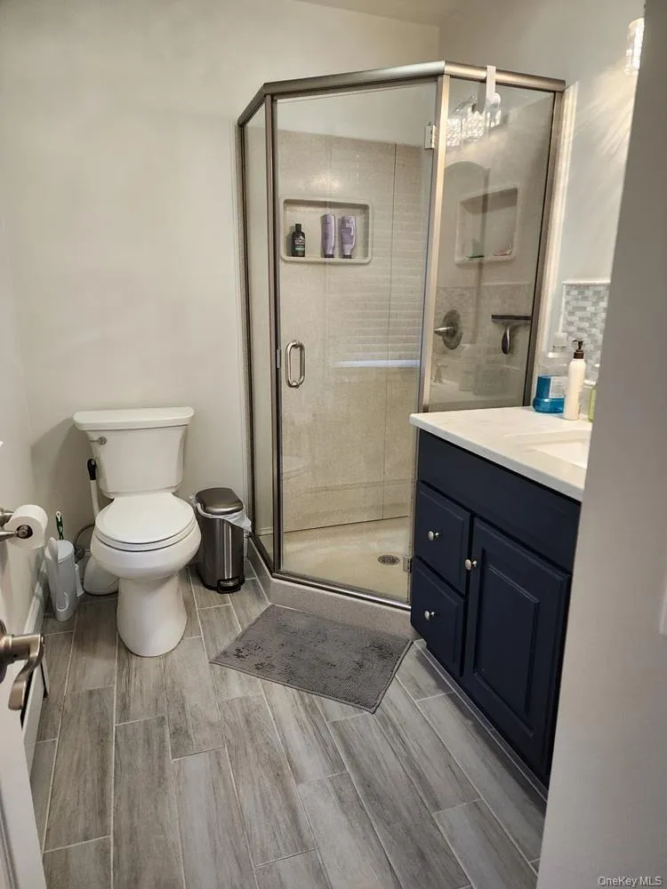 Bathroom featuring a corner glass-enclosed shower with integrated shelving, a dark blue vanity with a white countertop and under mount sink, and wood-finish tile flooring Bathroom featuring a corner glass-enclosed shower with integrated shelving, a dark blue vanity with a white countertop and under mount sink, and wood-finish tile flooring