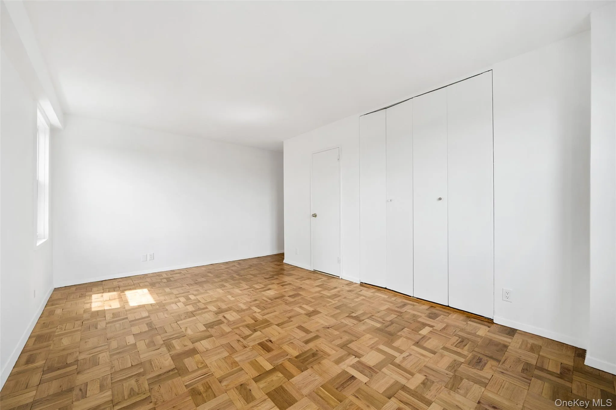 Bright room featuring parquet wood flooring, white walls, a large window, a full wall of bi-fold closet doors, and a separate interior door Bright room featuring parquet wood flooring, white walls, a large window, a full wall of bi-fold closet doors, and a separate interior door