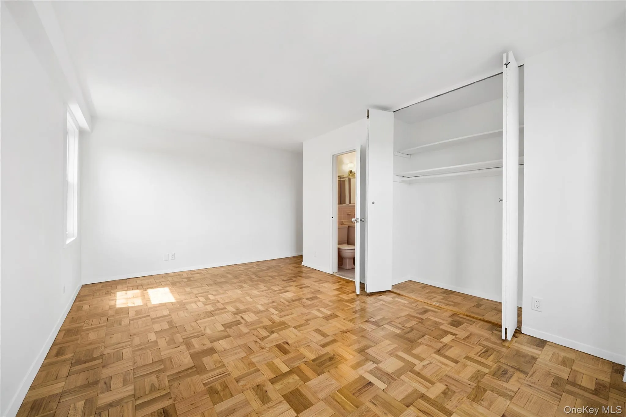Room featuring parquet wood flooring, white painted walls, a single window, and a bi-fold door closet with shelving and hanging rods Room featuring parquet wood flooring, white painted walls, a single window, and a bi-fold door closet with shelving and hanging rods