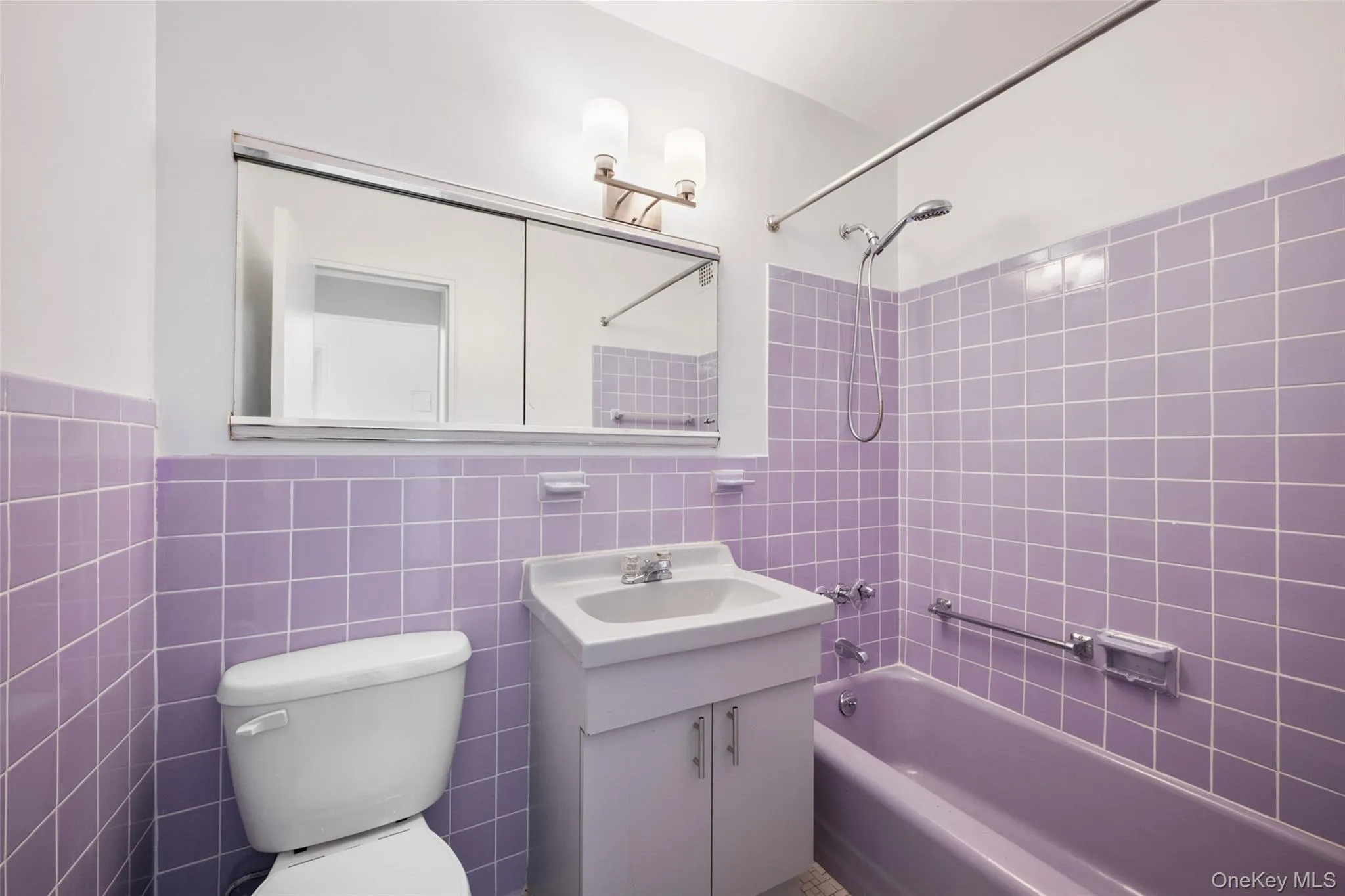 Main bathroom featuring purple square tile walls, a matching purple bathtub, a white vanity with an integrated sink, a mirrored medicine cabinet, and a two-bulb wall sconce Main bathroom featuring purple square tile walls, a matching purple bathtub, a white vanity with an integrated sink, a mirrored medicine cabinet, and a two-bulb wall sconce