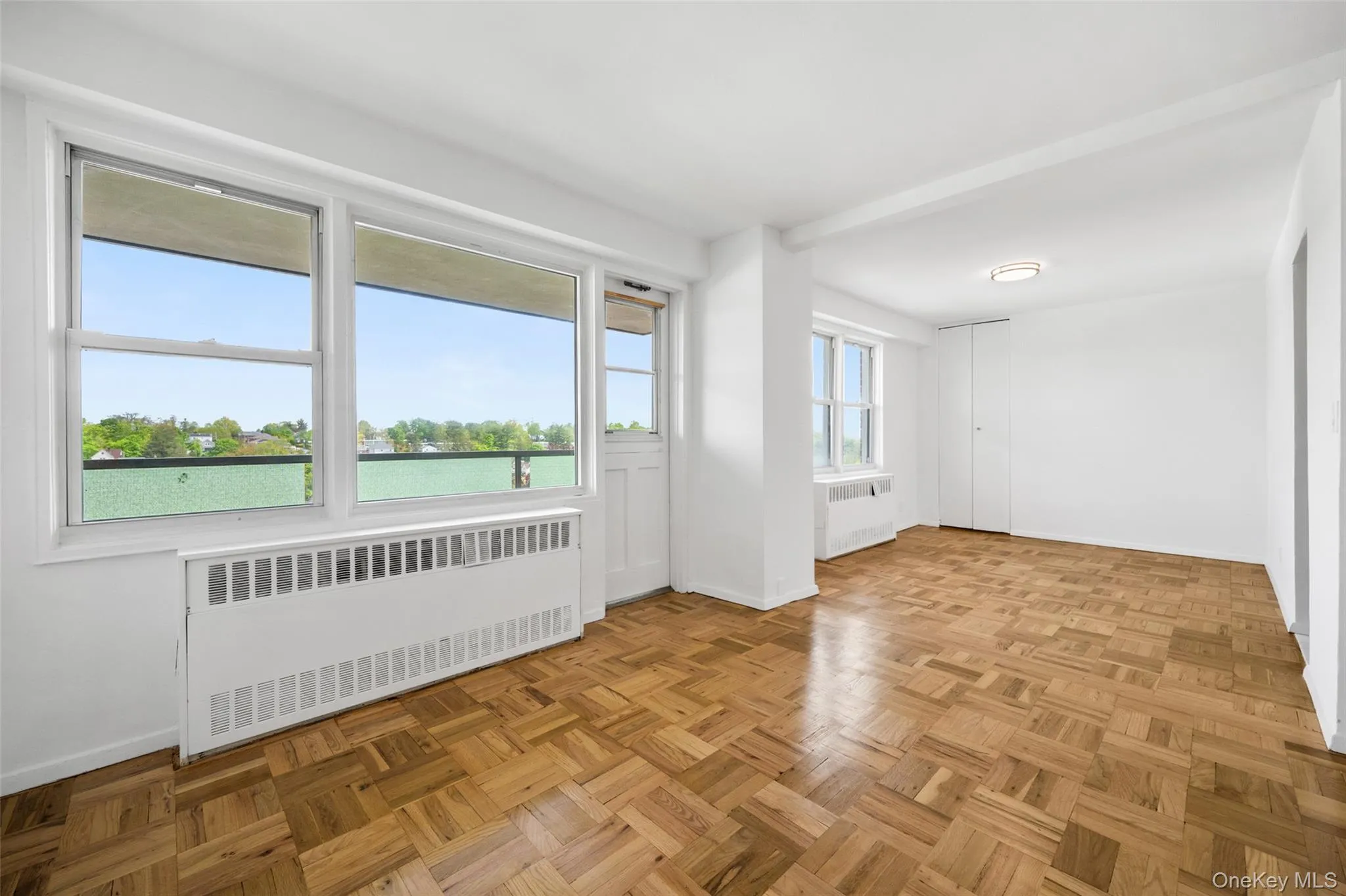 Sun-drenched interior featuring expansive windows, parquet wood-finish flooring, white painted walls, and an integrated radiator unit Sun-drenched interior featuring expansive windows, parquet wood-finish flooring, white painted walls, and an integrated radiator unit
