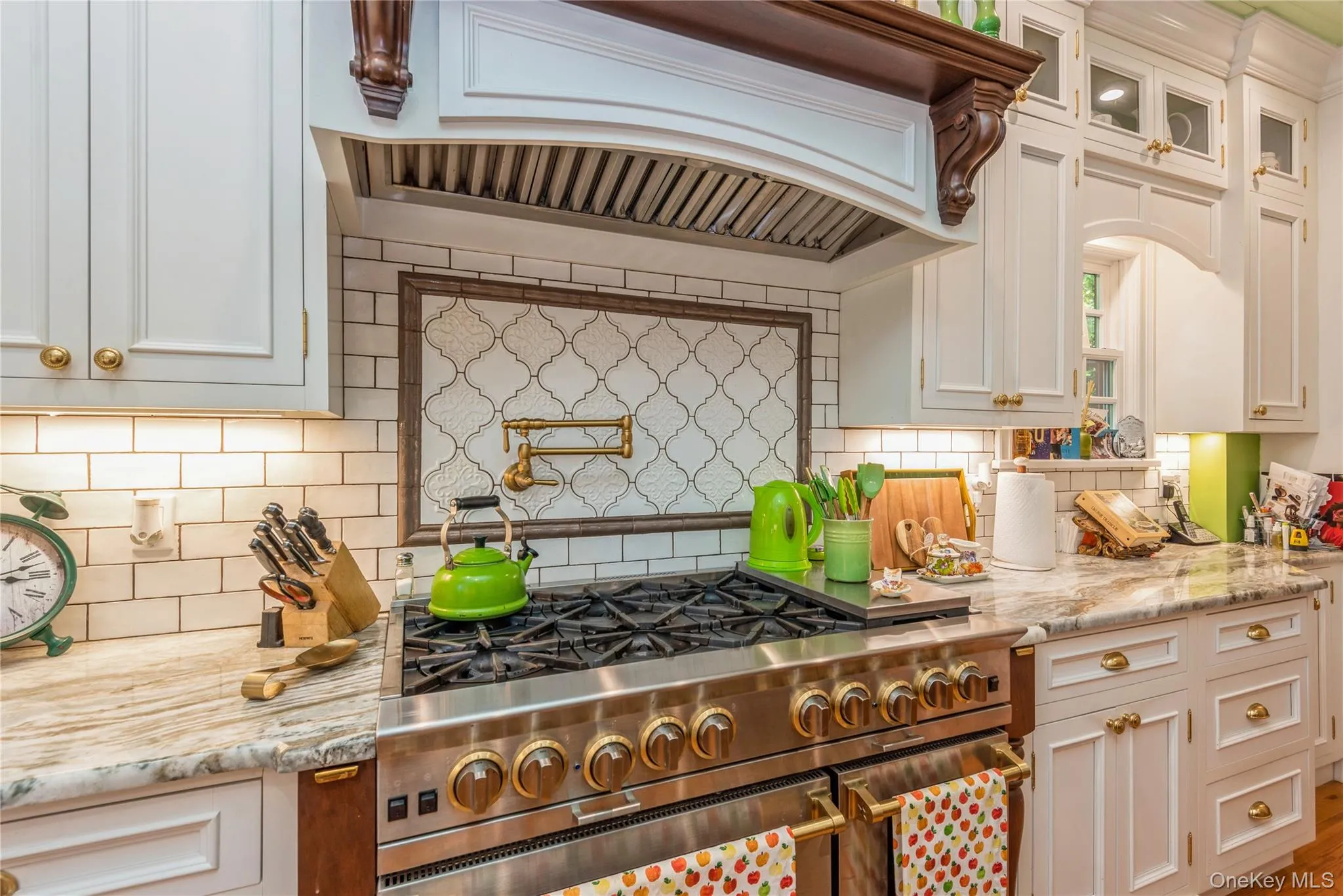 Professional-grade stainless steel range with brass-tone accents, set against a subway tile backsplash featuring an arabesque-patterned inset and a wall-mounted pot filler Professional-grade stainless steel range with brass-tone accents, set against a subway tile backsplash featuring an arabesque-patterned inset and a wall-mounted pot filler