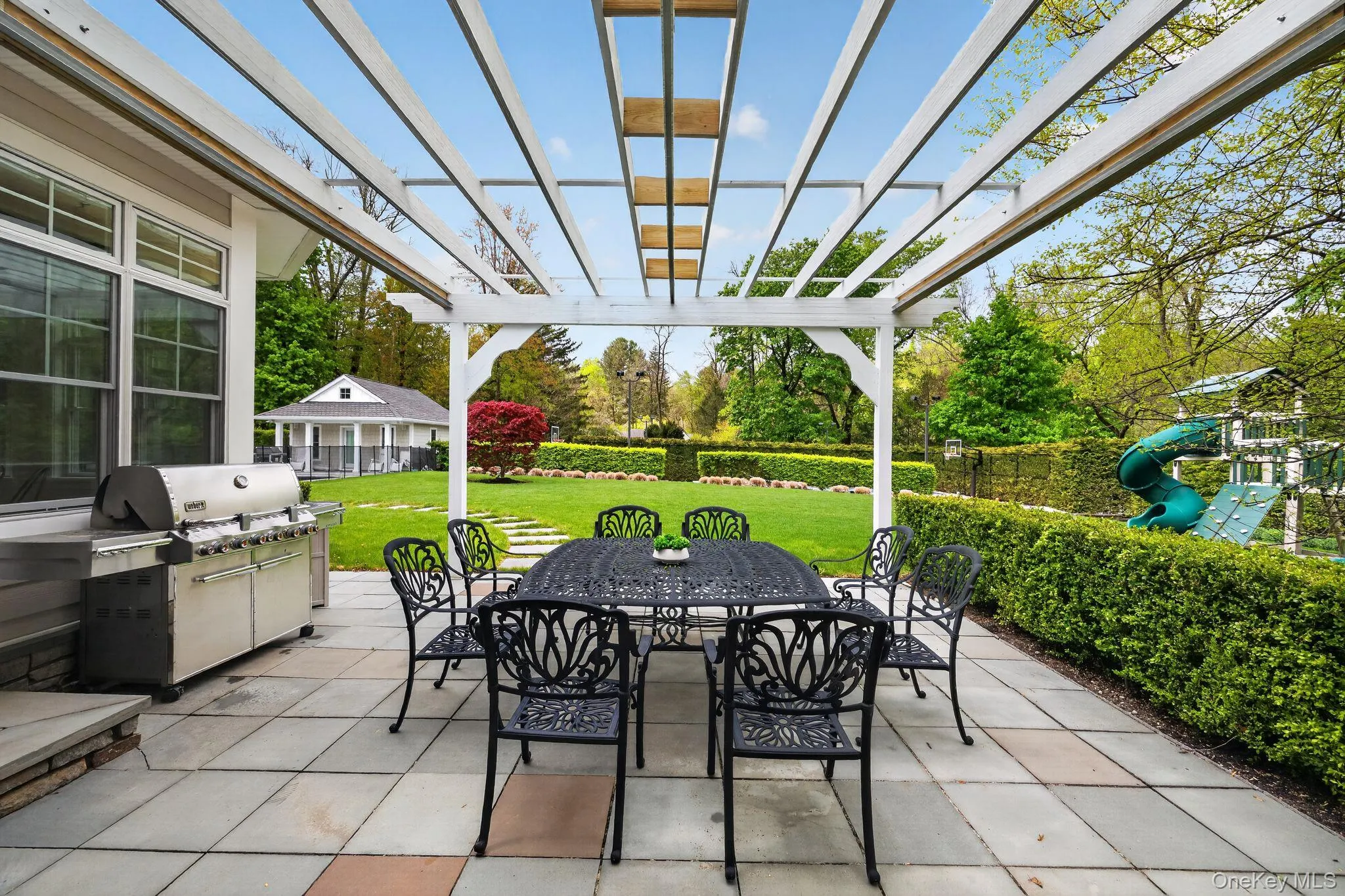 Expansive stone patio featuring a white pergola, outdoor grill, and manicured hedges Expansive stone patio featuring a white pergola, outdoor grill, and manicured hedges