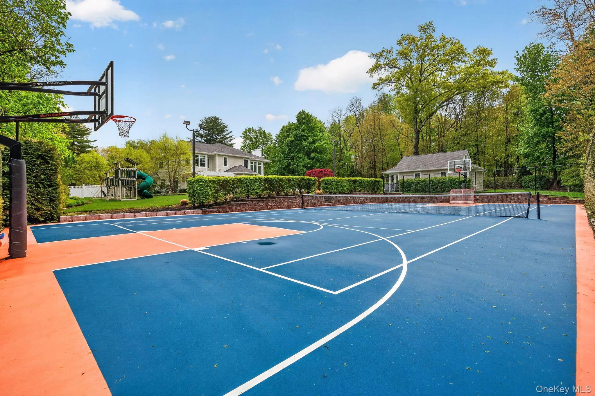 Multipurpose sports court featuring blue and orange surfacing with white line markings, basketball hoop, and tennis net Multipurpose sports court featuring blue and orange surfacing with white line markings, basketball hoop, and tennis net
