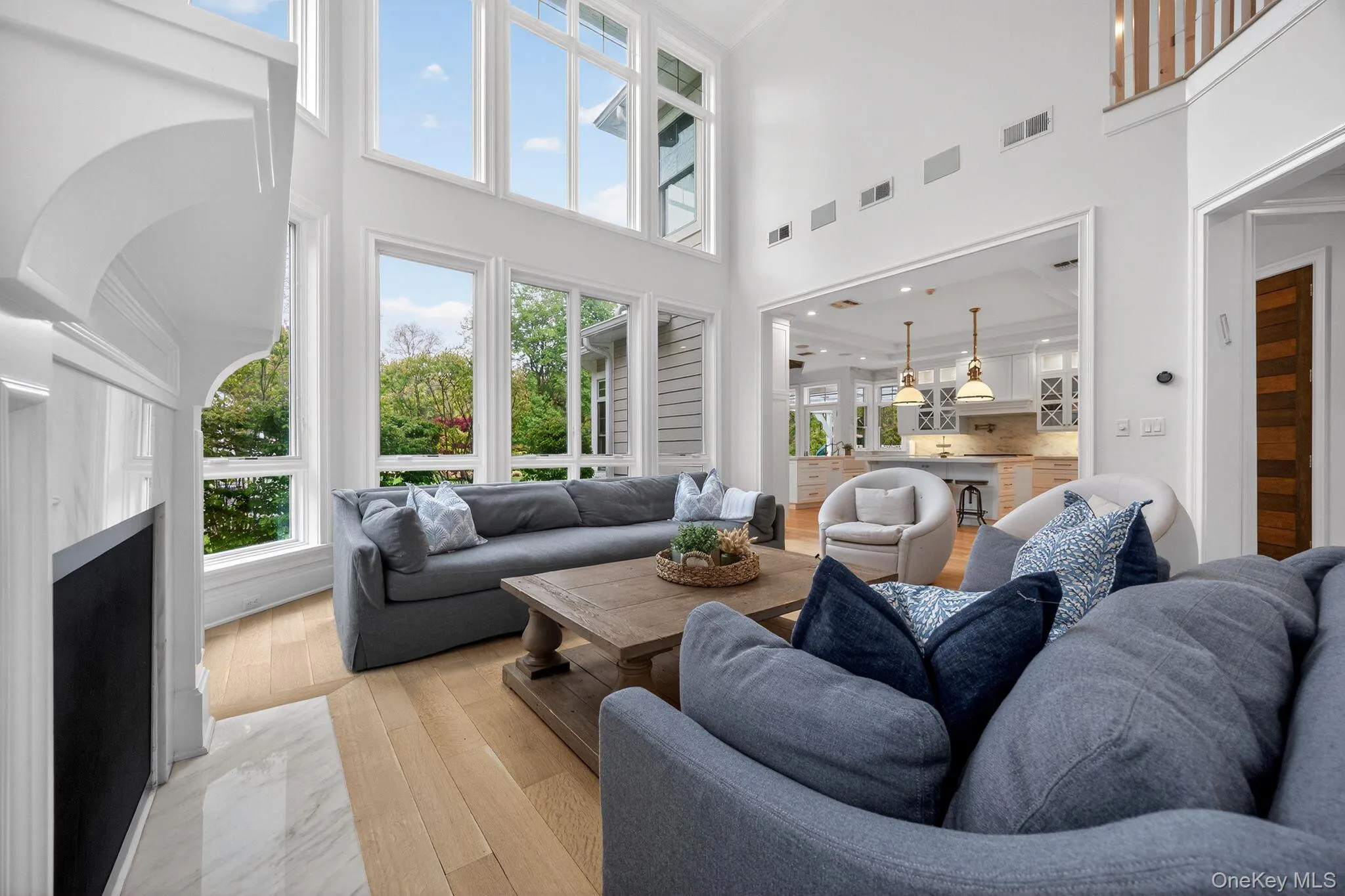 Two-story living area featuring a fireplace with a marble-style hearth, abundant natural light from numerous windows, light wood-finish flooring, and an open concept layout to the kitchen Two-story living area featuring a fireplace with a marble-style hearth, abundant natural light from numerous windows, light wood-finish flooring, and an open concept layout to the kitchen