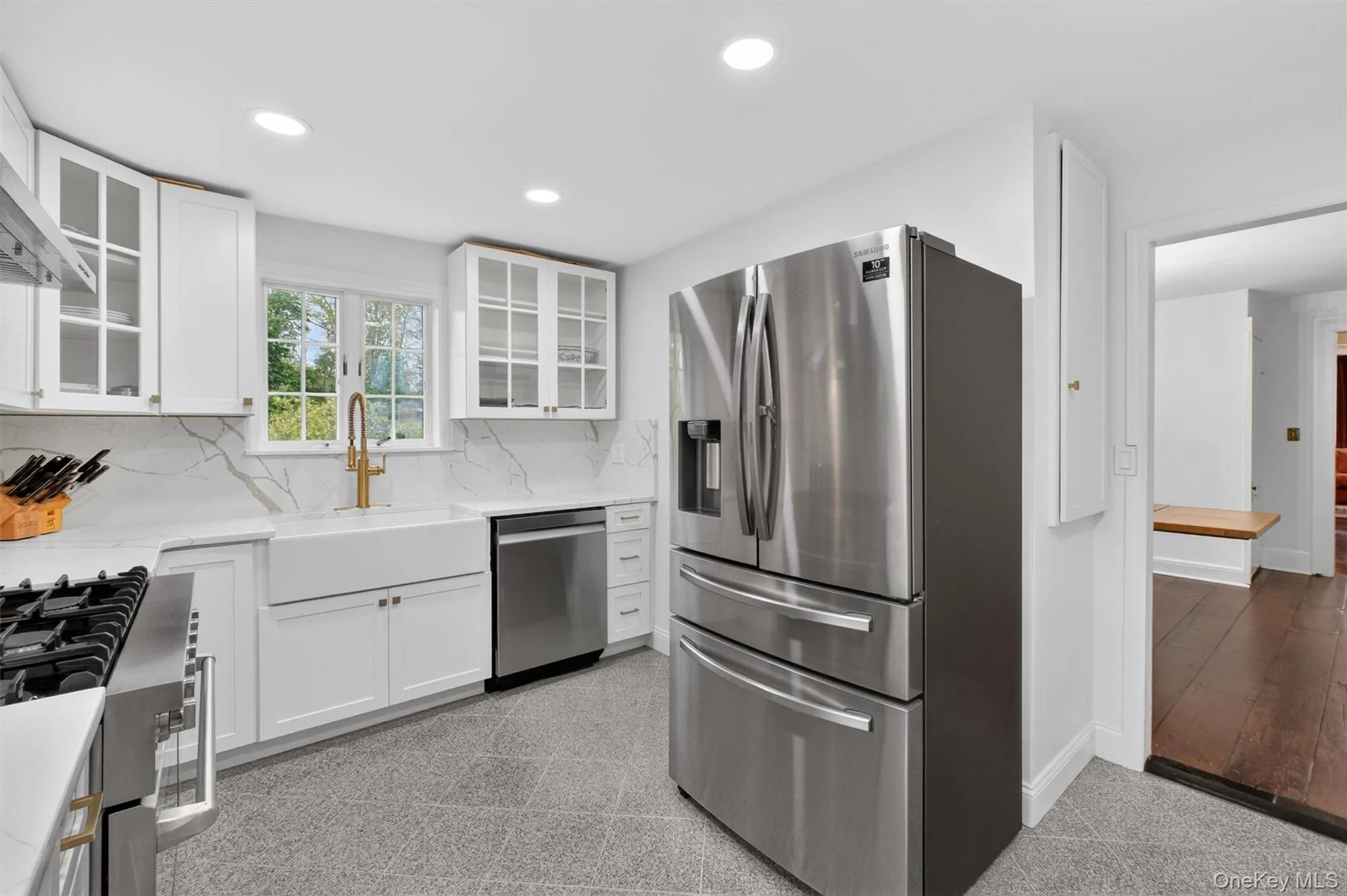 Bright kitchen featuring white cabinetry, white quartz countertops with veining, a stainless-steel range with hood, a white farmhouse sink, recessed lighting and gold ornamentation hardware Bright kitchen featuring white cabinetry, white quartz countertops with veining, a stainless-steel range with hood, a white farmhouse sink, recessed lighting and gold ornamentation hardware