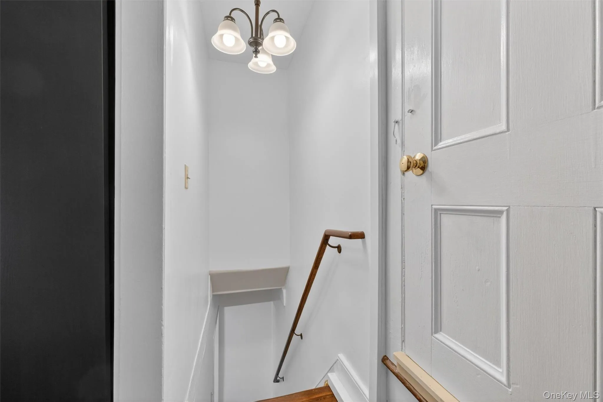 Stairwell featuring a traditional three-light chandelier, a wood-finish handrail, white wall paneling, and a white paneled door with a brass knob connecting to a side bedroom from the dining room side staircase. Stairwell featuring a traditional three-light chandelier, a wood-finish handrail, white wall paneling, and a white paneled door with a brass knob connecting to a side bedroom from the dining room side staircase.
