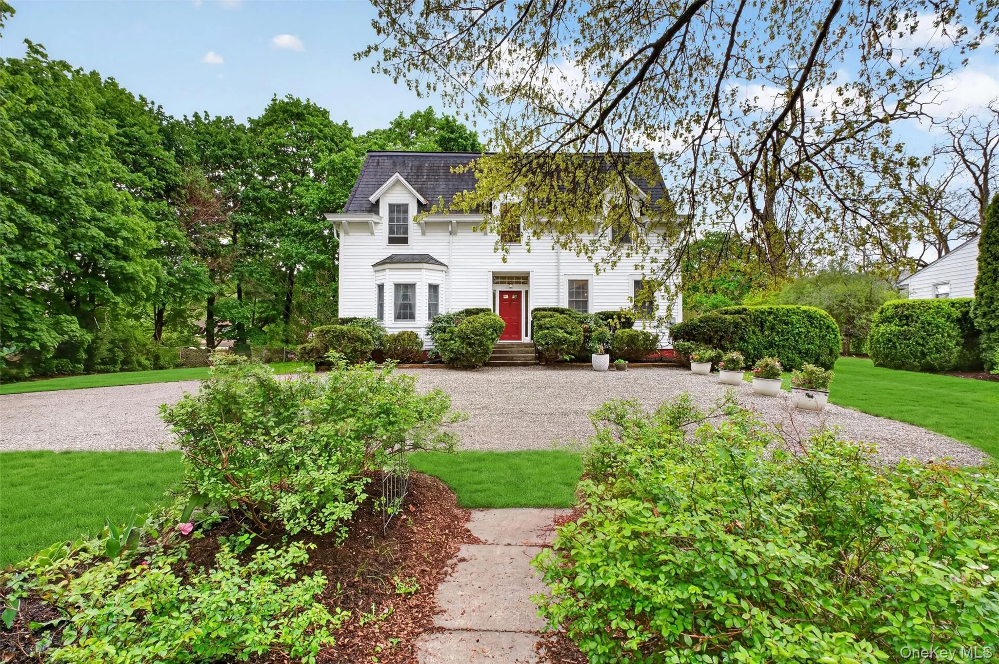 Exterior of colonial residence with red door and mansard roof. Gravel driveway with manicured gardens surrounding. Exterior of colonial residence with red door and mansard roof. Gravel driveway with manicured gardens surrounding.