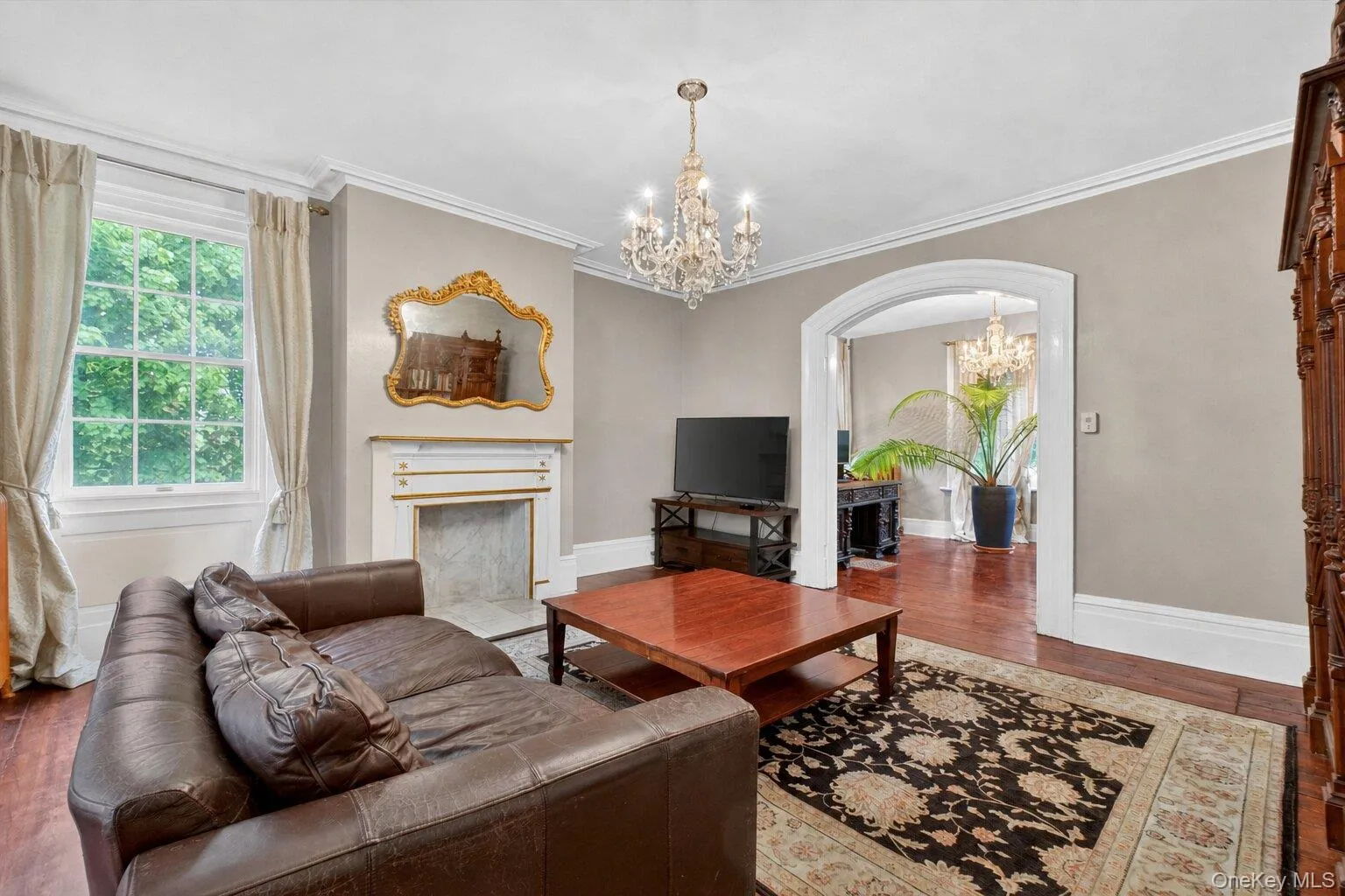 Living area featuring crystal chandelier, hardwood floors, a decorative fireplace with a mantel, an arched doorway leading to another living area, and white trim molding Living area featuring crystal chandelier, hardwood floors, a decorative fireplace with a mantel, an arched doorway leading to another living area, and white trim molding
