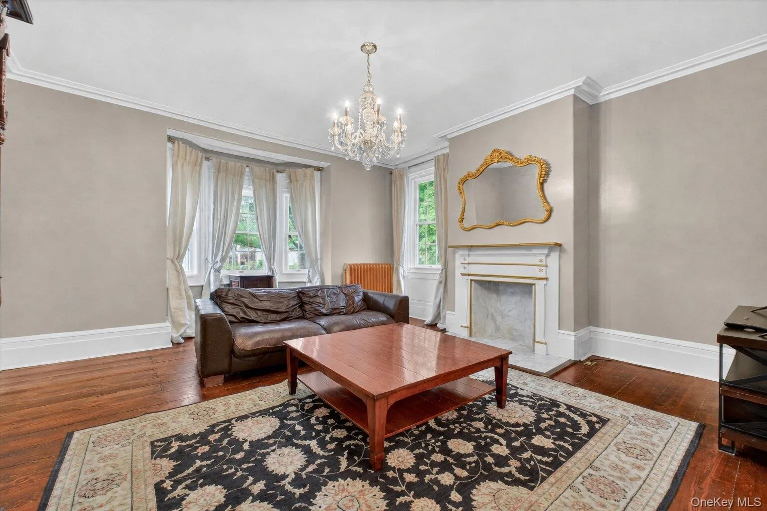 Living area featuring crystal chandelier, hardwood floors, a decorative fireplace with a mantel, bay window looking onto the lawn, and white trim molding Living area featuring crystal chandelier, hardwood floors, a decorative fireplace with a mantel, bay window looking onto the lawn, and white trim molding