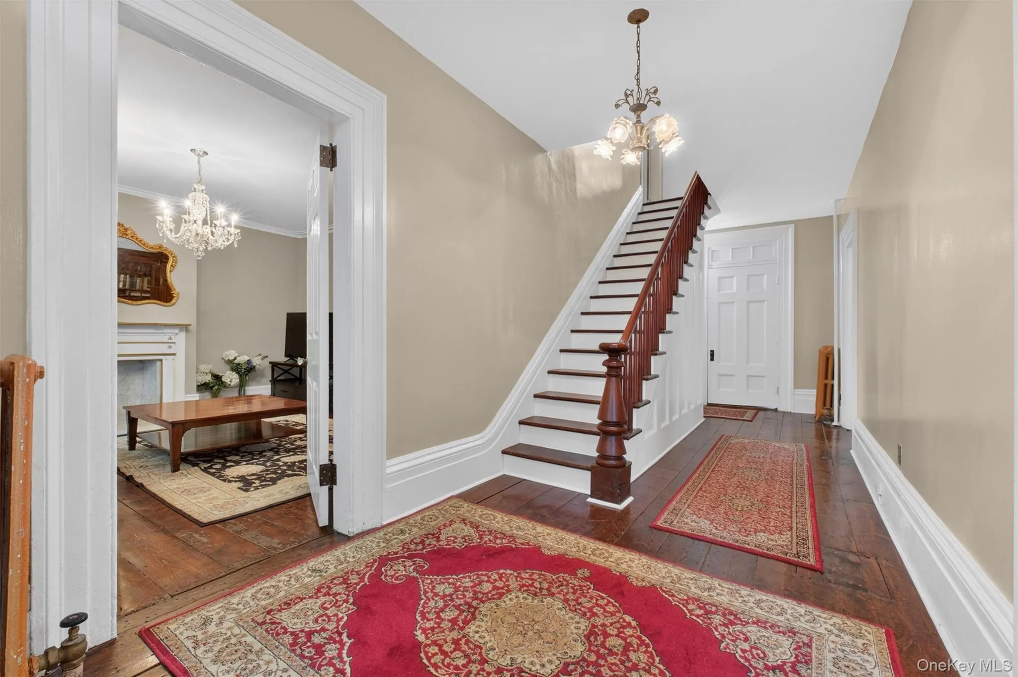 Foyer showcasing hardwood flooring, a classic chandelier, a dark wood staircase with balusters, hallway and door leading to a living area. Foyer showcasing hardwood flooring, a classic chandelier, a dark wood staircase with balusters, hallway and door leading to a living area.