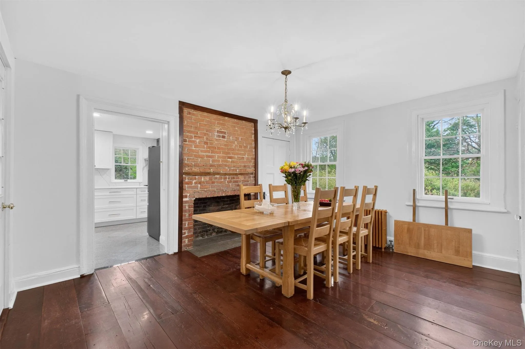 Dining room featuring a prominent exposed brick fireplace, hardwood flooring, large windows and a doorway leading to the kitchen. Dining room featuring a prominent exposed brick fireplace, hardwood flooring, large windows and a doorway leading to the kitchen.