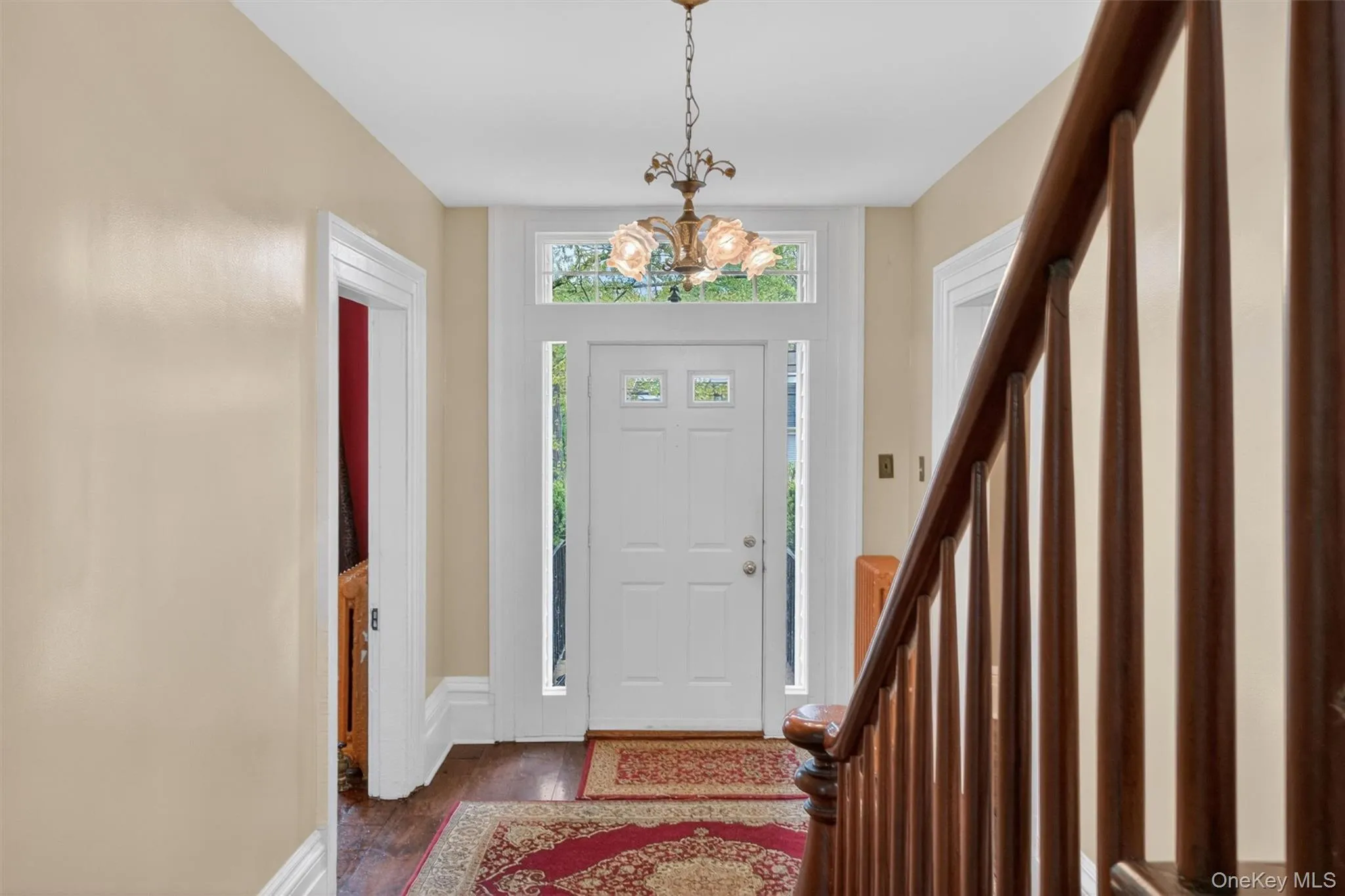 Foyer showcasing a white entry door with a transom window, hardwood flooring, a classic chandelier, and a dark wood staircase with balusters. Foyer showcasing a white entry door with a transom window, hardwood flooring, a classic chandelier, and a dark wood staircase with balusters.
