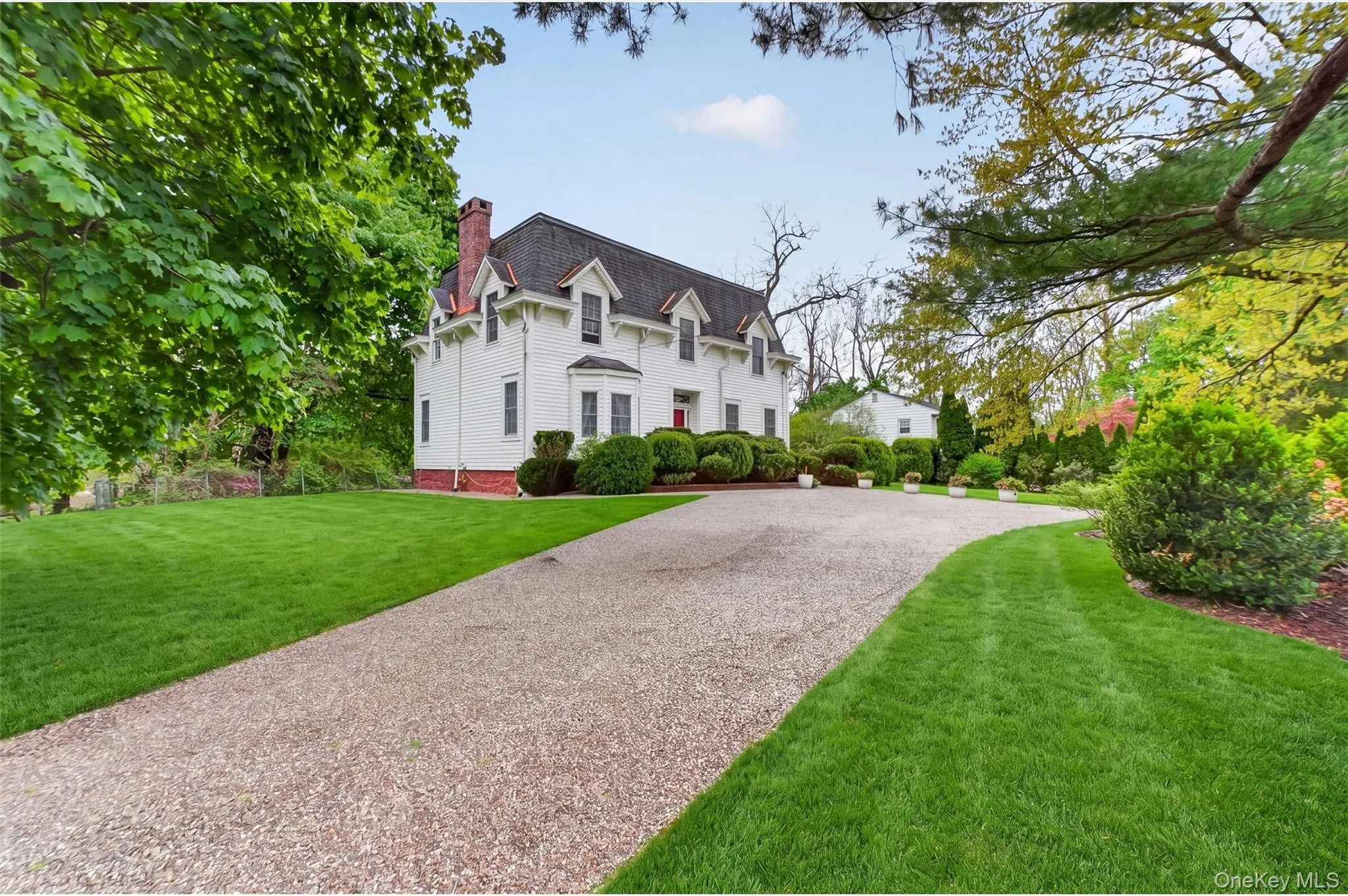Exterior of colonial residence with red door and mansard roof. Gravel driveway with manicured gardens and lawn surrounding. Exterior of colonial residence with red door and mansard roof. Gravel driveway with manicured gardens and lawn surrounding.