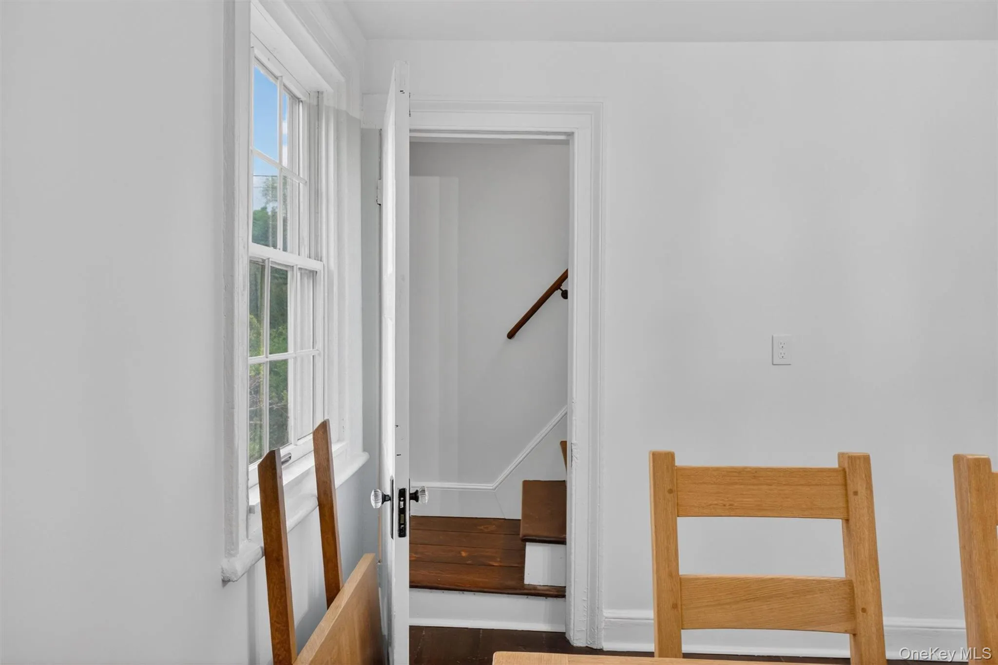 Bright interior with a double-hung window, white trim, and a doorway revealing a side staircase with wood-finish treads and a dark wood handrail. Bright interior with a double-hung window, white trim, and a doorway revealing a side staircase with wood-finish treads and a dark wood handrail.