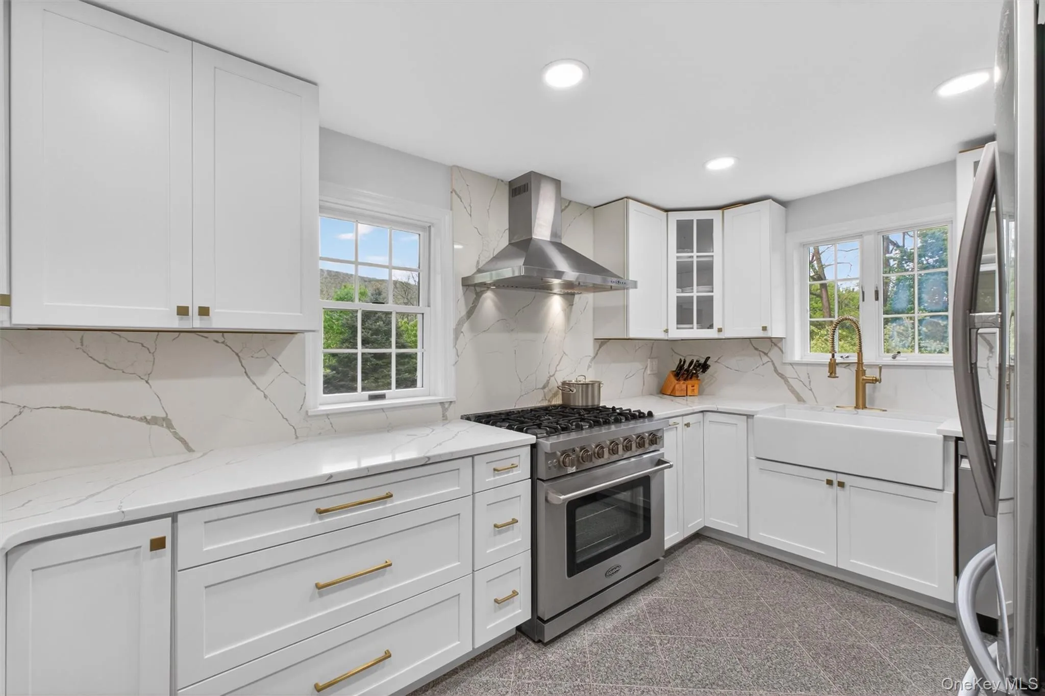 Bright kitchen featuring white cabinetry, white quartz countertops with veining, a stainless-steel range with hood, a white farmhouse sink, recessed lighting and gold ornamentation hardware Bright kitchen featuring white cabinetry, white quartz countertops with veining, a stainless-steel range with hood, a white farmhouse sink, recessed lighting and gold ornamentation hardware