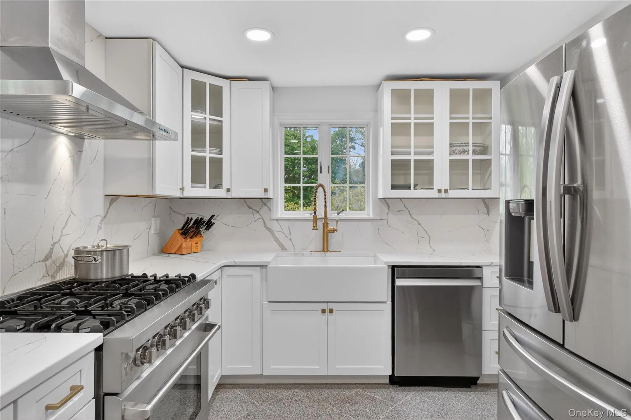 Bright kitchen featuring white cabinetry, white quartz countertops with veining, a stainless-steel range with hood, a white farmhouse sink, recessed lighting and gold ornamentation hardware Bright kitchen featuring white cabinetry, white quartz countertops with veining, a stainless-steel range with hood, a white farmhouse sink, recessed lighting and gold ornamentation hardware