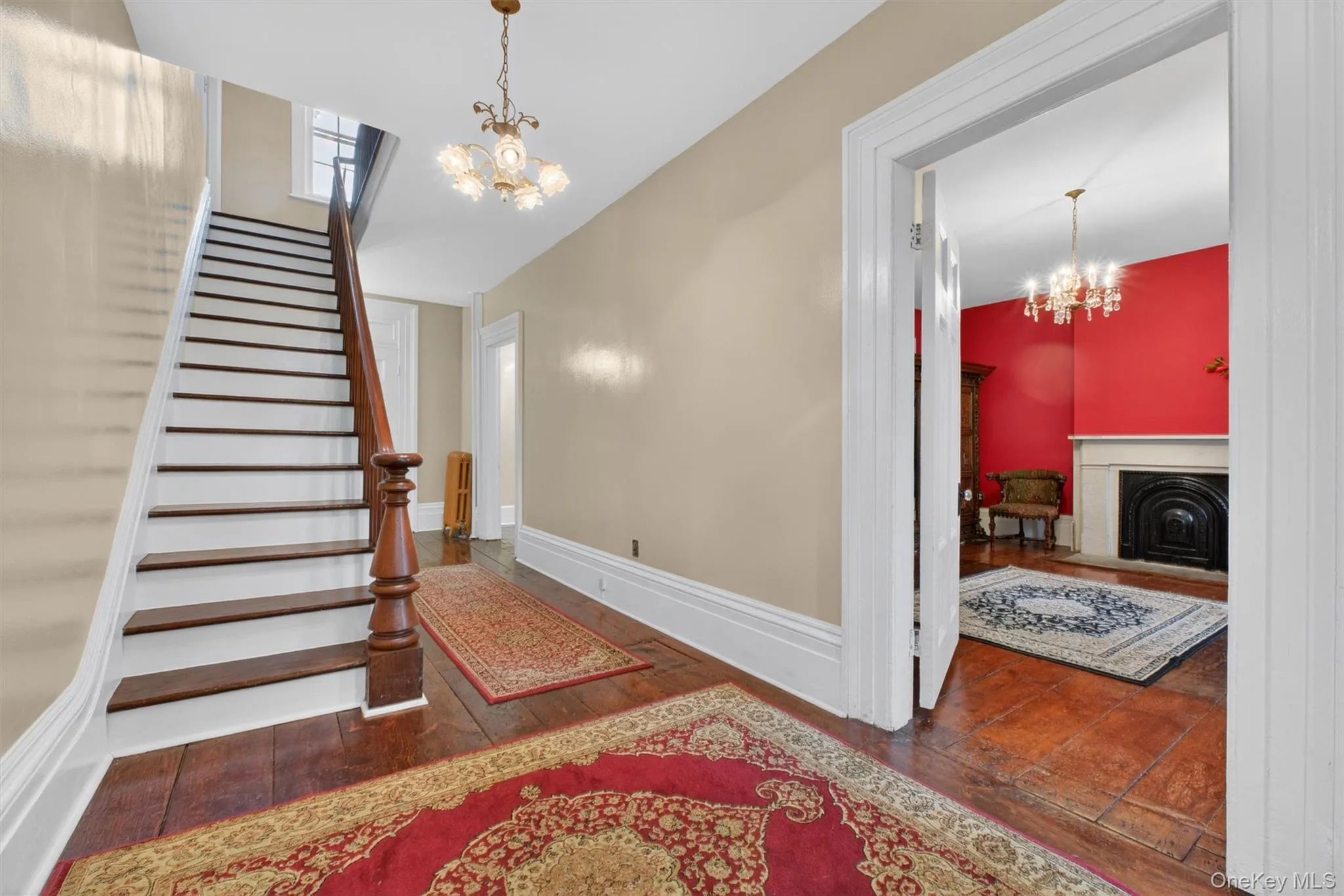 Foyer showcasing hardwood flooring, a classic chandelier, a dark wood staircase with balusters, hallway and door leading to a living area. Foyer showcasing hardwood flooring, a classic chandelier, a dark wood staircase with balusters, hallway and door leading to a living area.